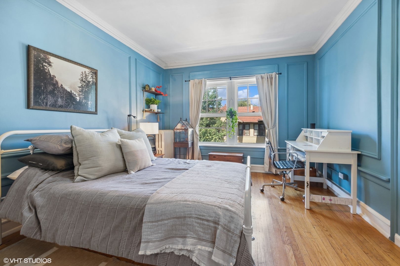 Bedroom with blue paneled walls, gray bedding, hardwood floors, and a white desk beside the window.