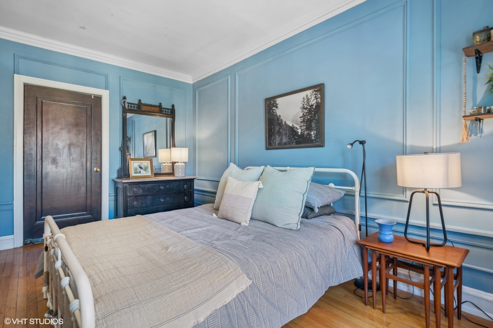 Bedroom with blue paneled walls, a white metal bed, layered neutral bedding, and hardwood floors.