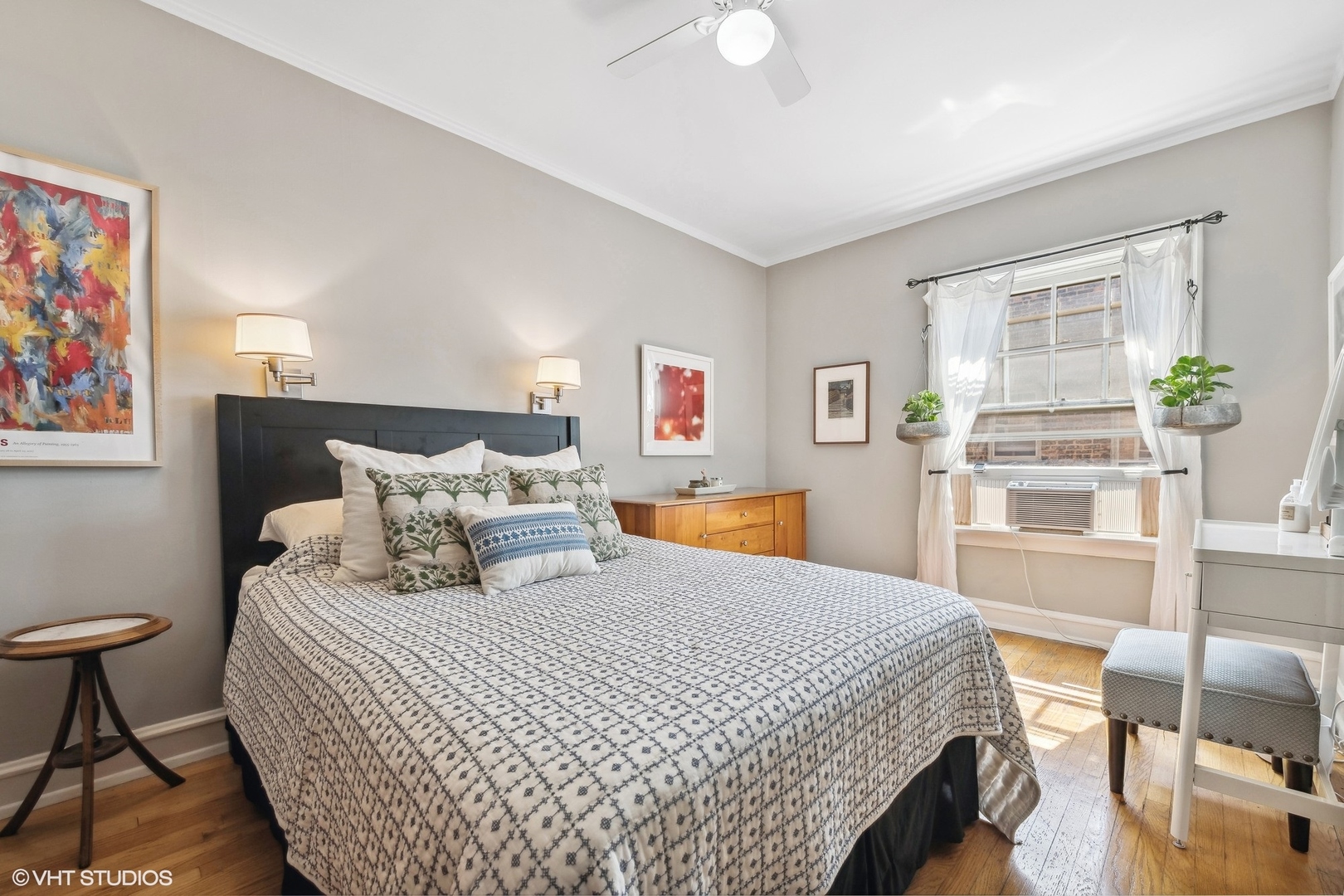 Furnished bedroom with a patterned bedspread, hardwood floors, and a bright window with sheer curtains.