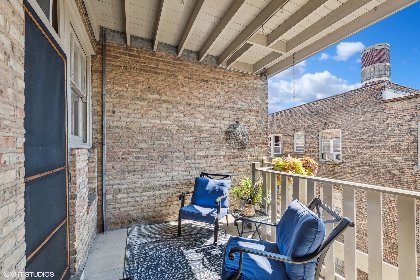 Covered brick balcony with blue-cushioned chairs, potted plants, and views of neighboring buildings.