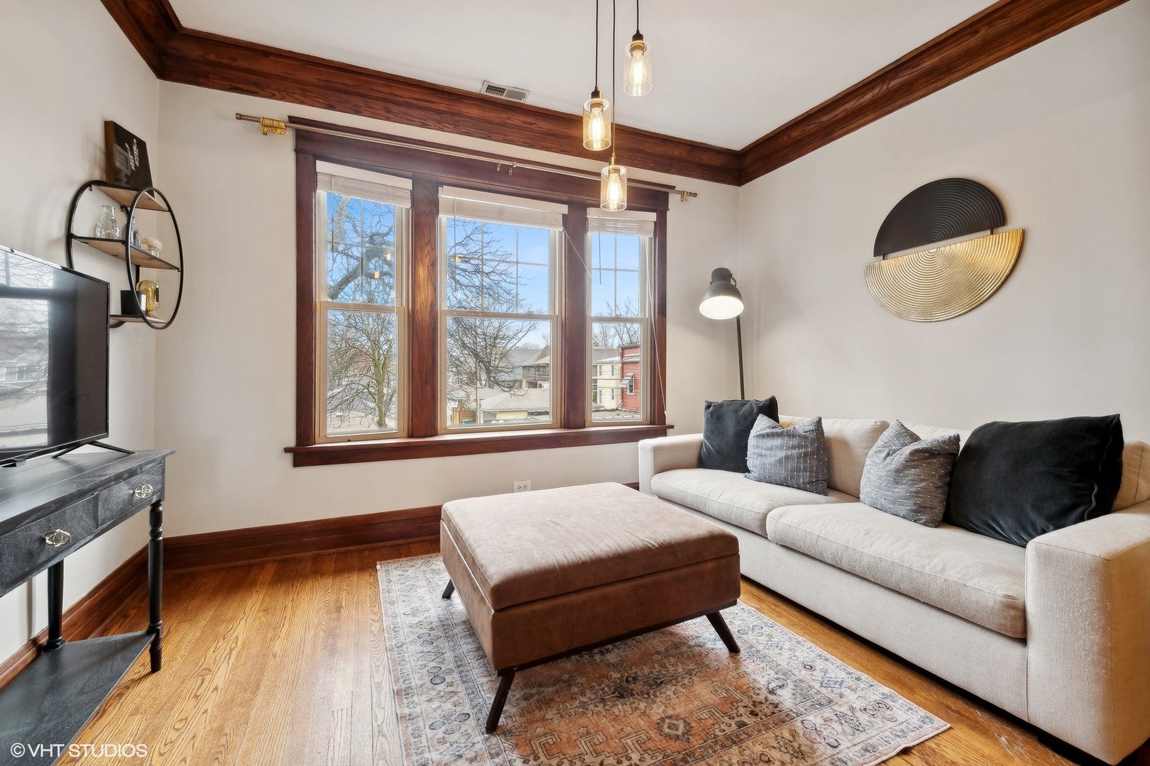 Living room with a cream sectional, patterned rug, hardwood floors, and three large wood-trimmed windows.