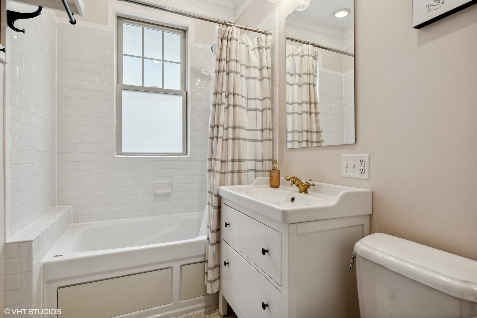 Bathroom with white tiled tub, window, striped shower curtain, and vanity with brass faucet.
