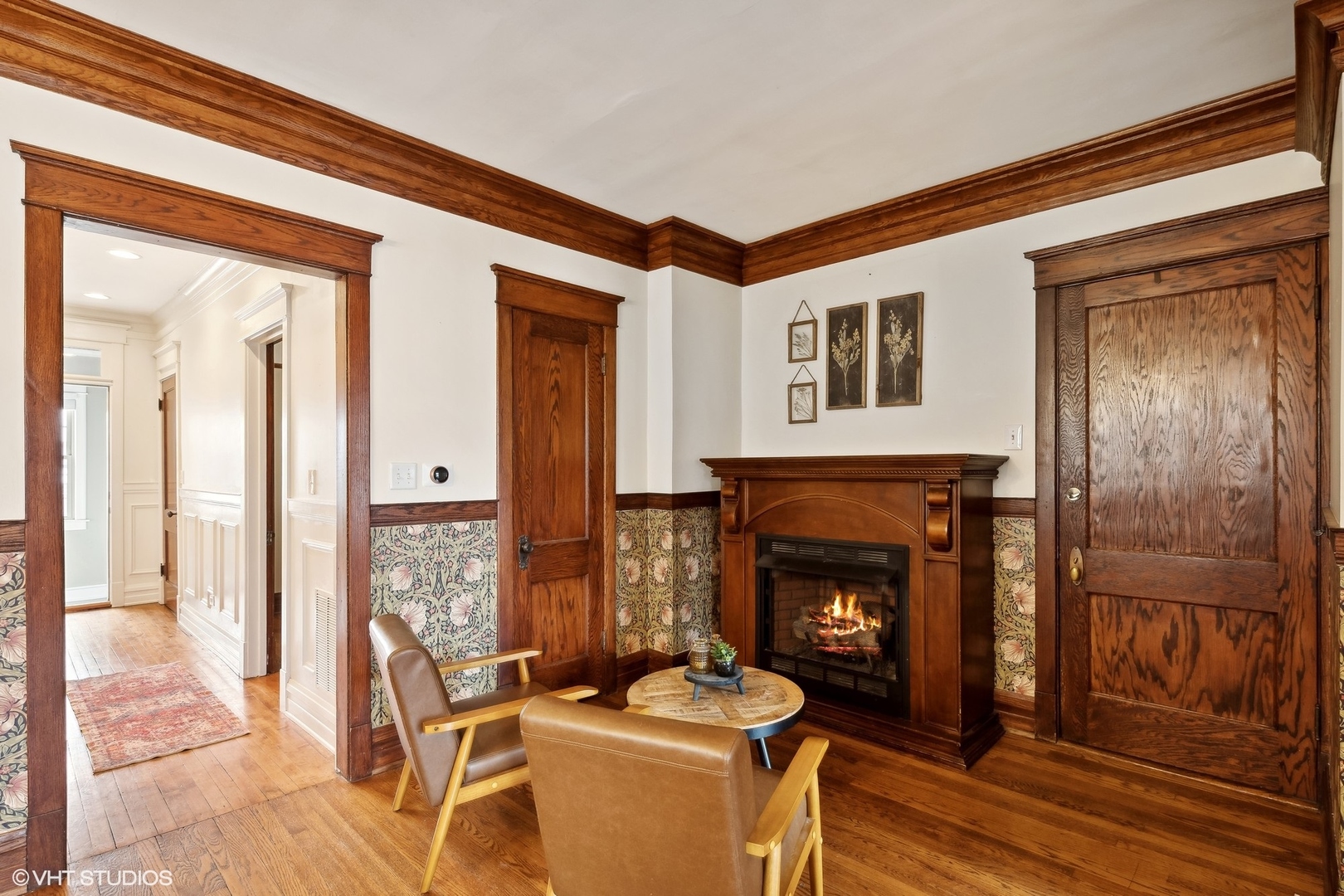 Living room with a lit fireplace, rich wood trim, floral wall covering, and two leather chairs.