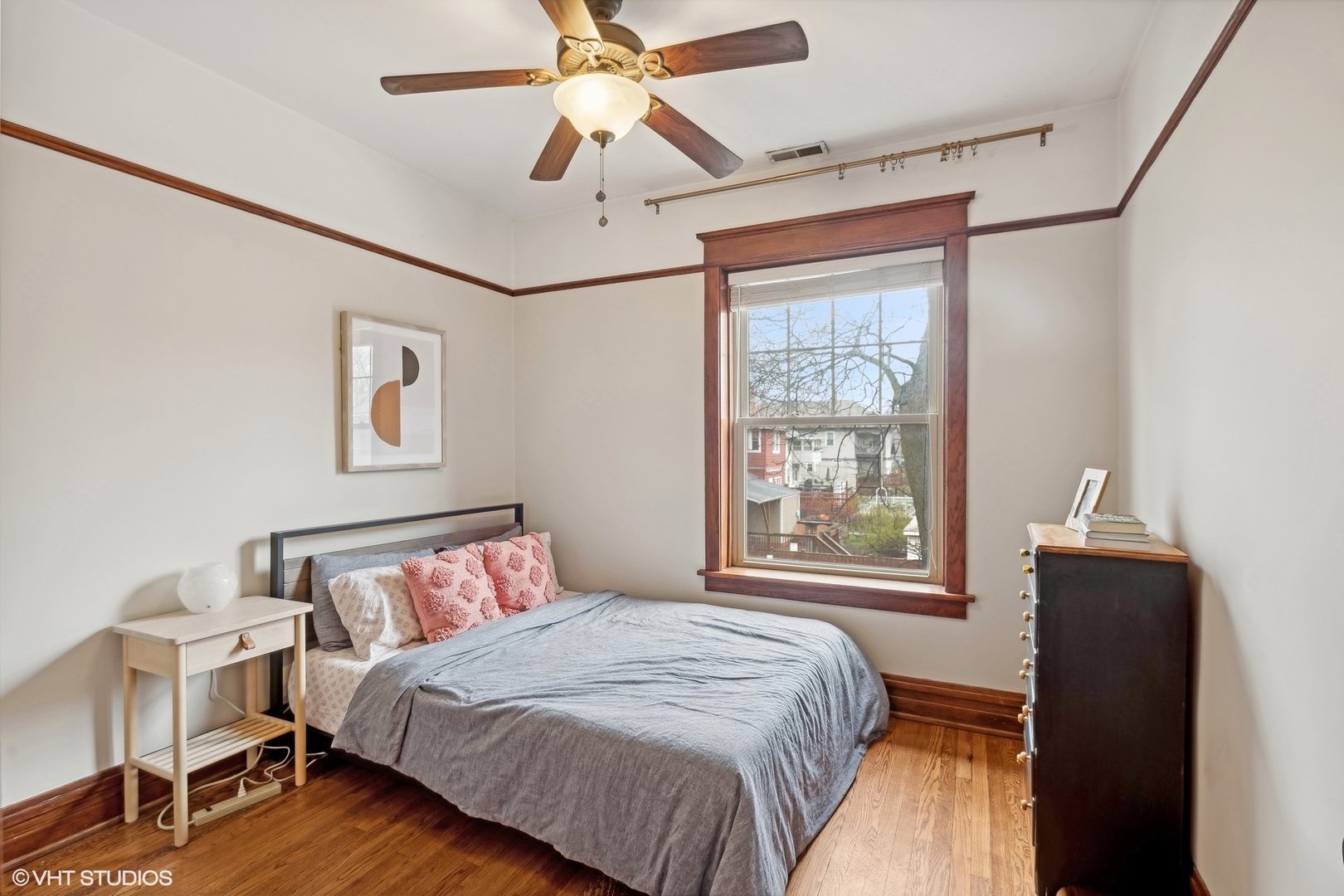 Bedroom with gray bedding, hardwood floors, dark wood trim, and a large sunlit window.