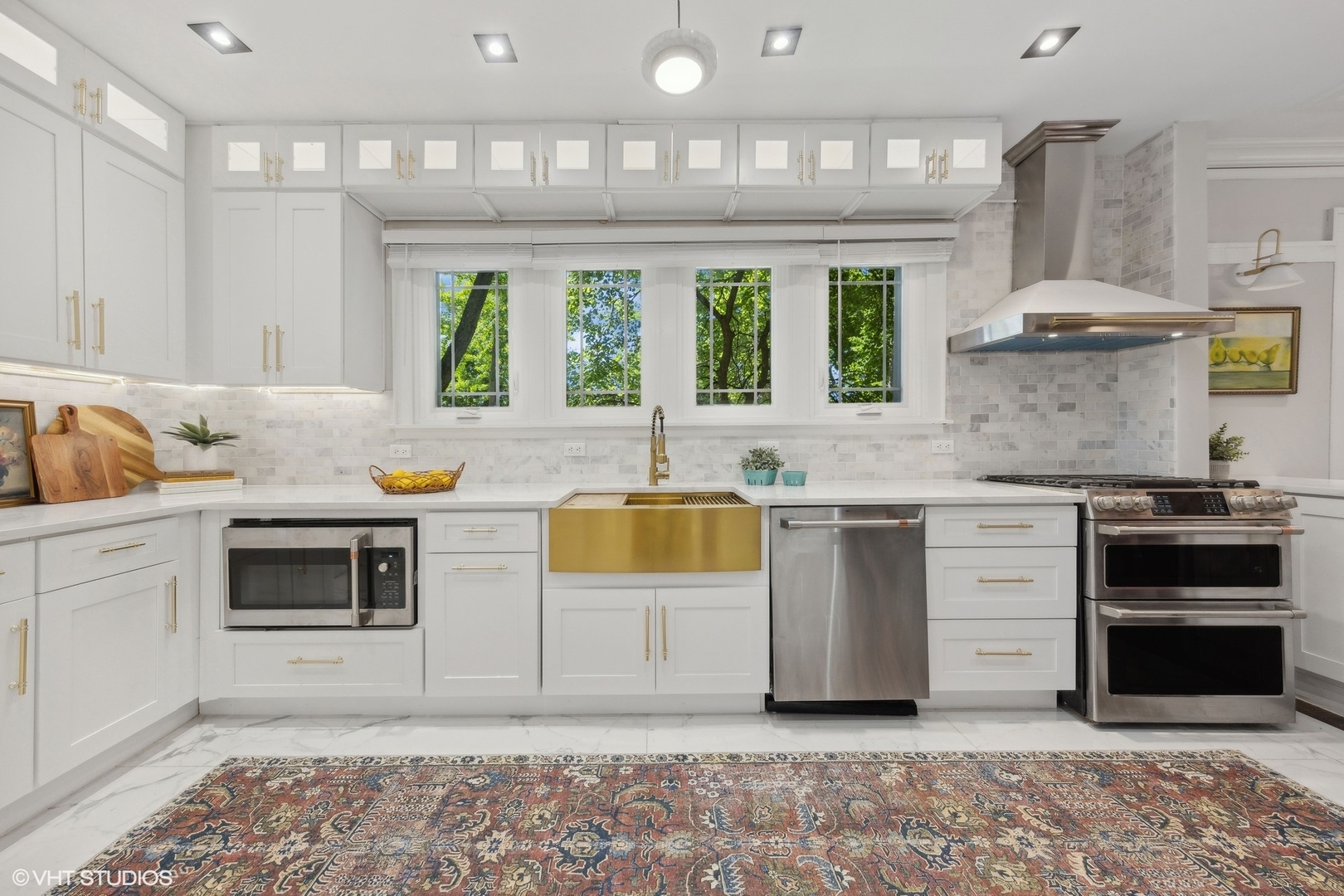 Bright white kitchen with brass farmhouse sink, stainless-steel appliances, and windows overlooking leafy trees.