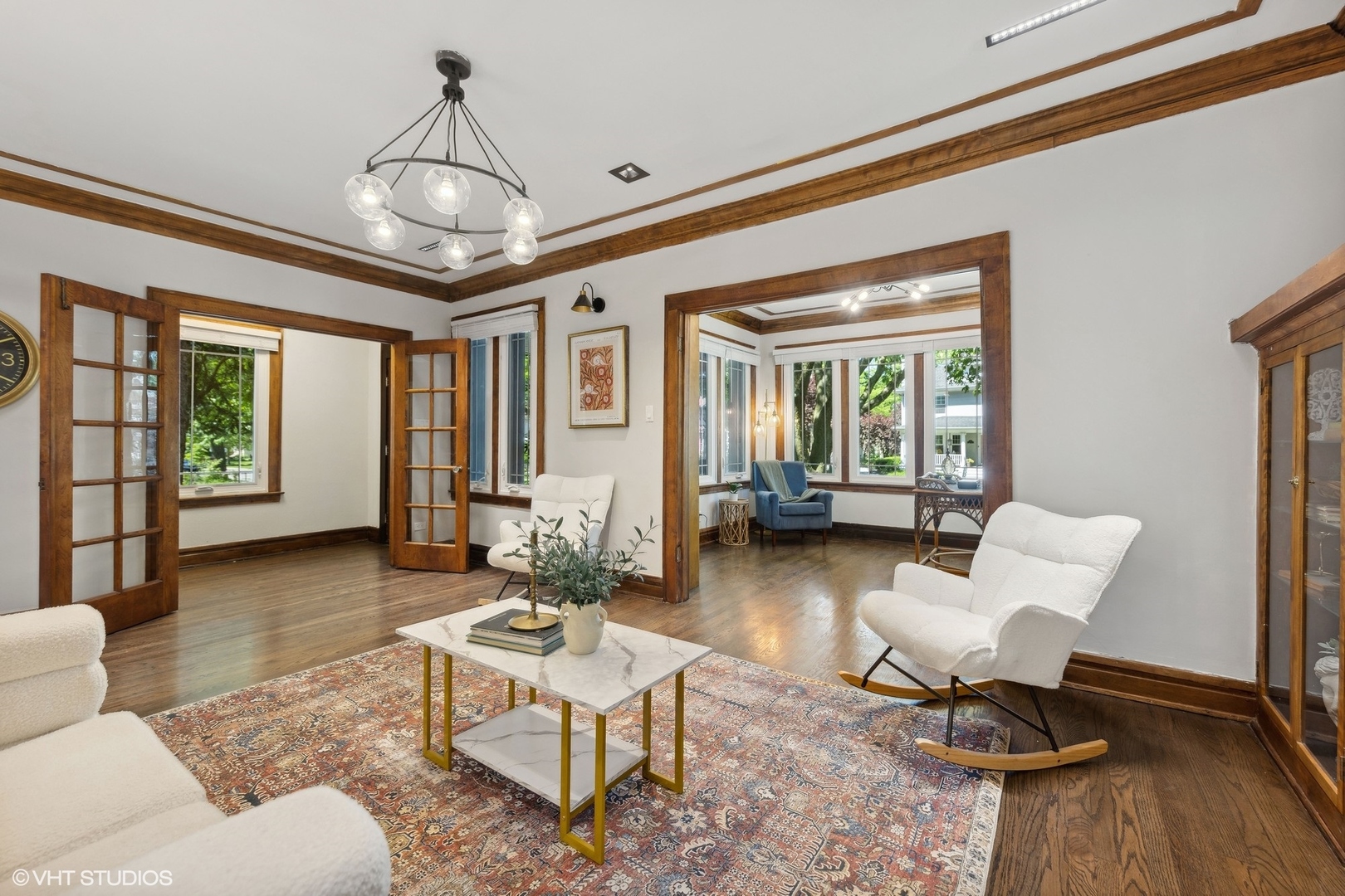 Living room with wood-trimmed French doors, hardwood floors, patterned rug, and a bright adjoining sitting area.