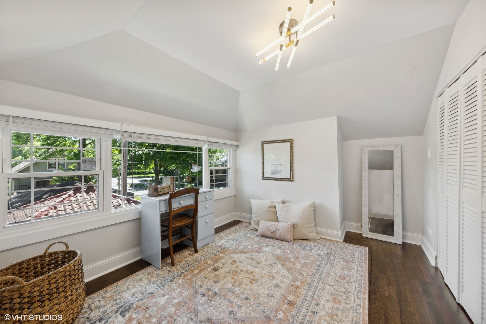 Bright room with large windows, a small desk, louvered closet doors, and a patterned rug.