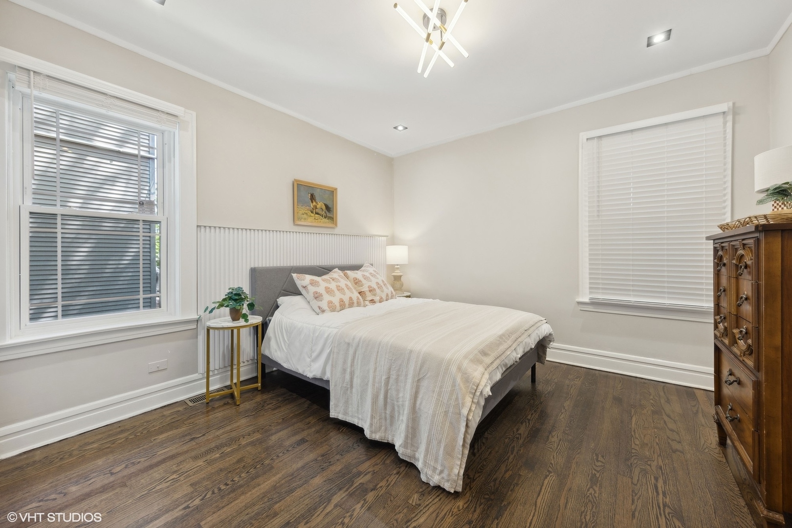 Furnished bedroom with a white-covered bed, dark hardwood floors, and windows with blinds.