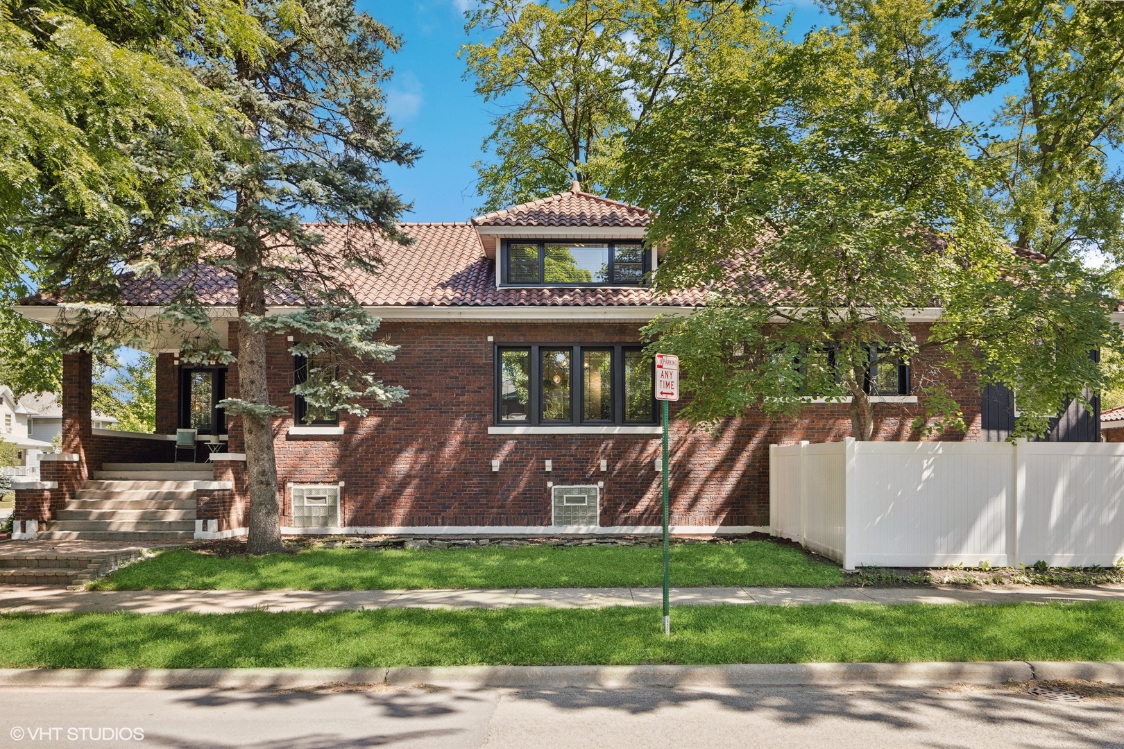 Brick home with a red tile roof, shaded by mature trees and bordered by a white fence.