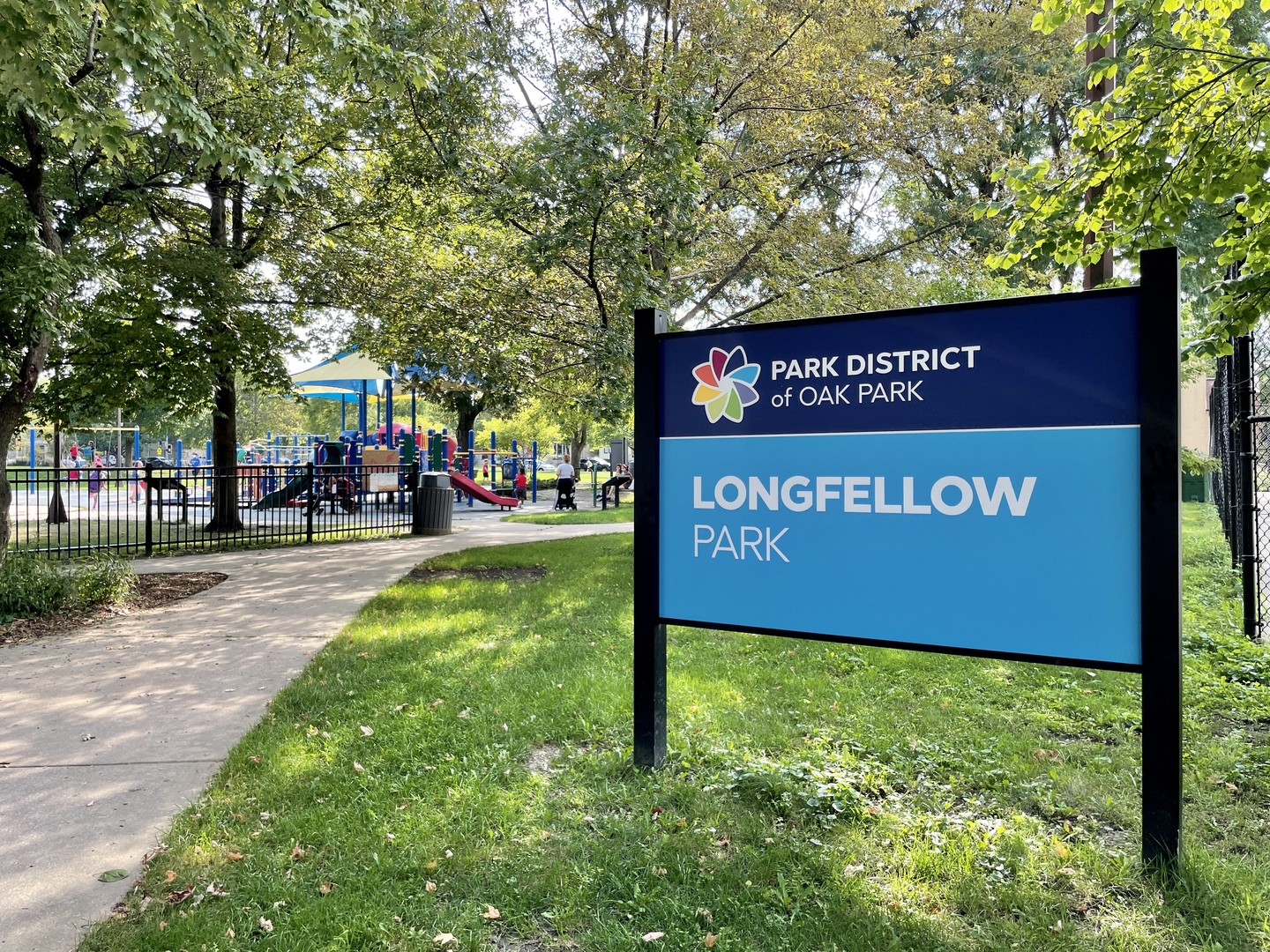 Longfellow Park sign beside a tree-shaded playground and paved path.