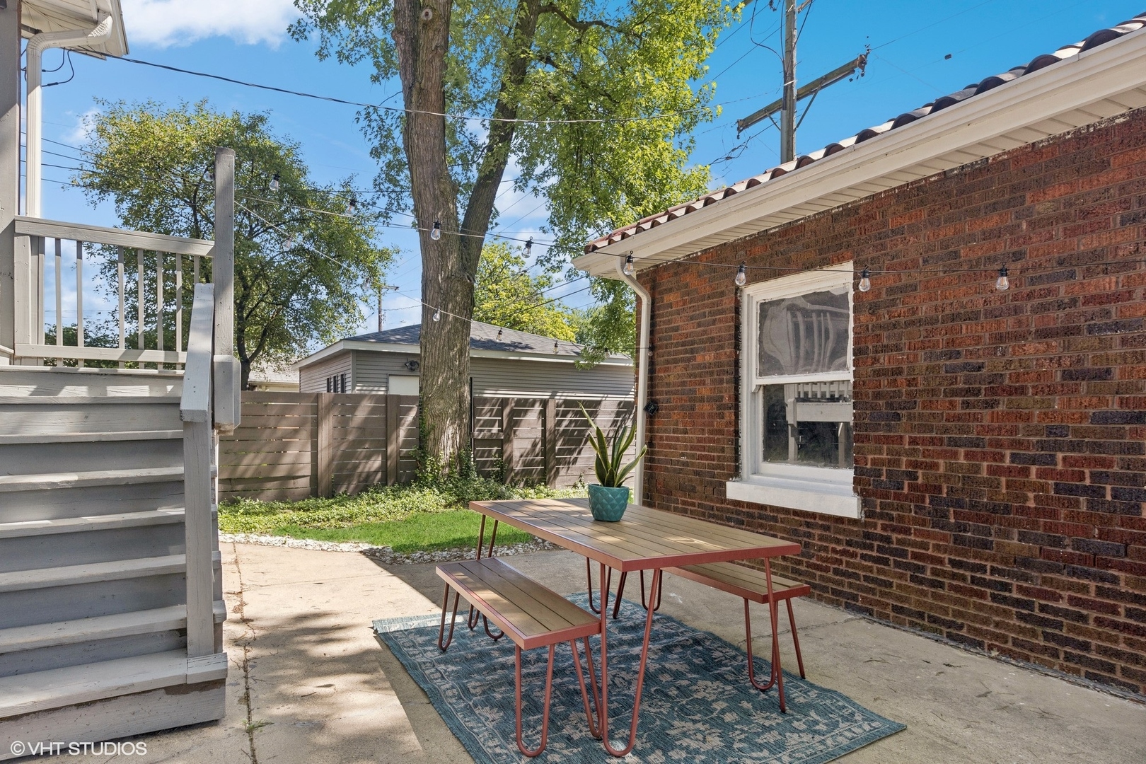 Backyard patio with a picnic table and benches beside a red brick wall and white deck steps.