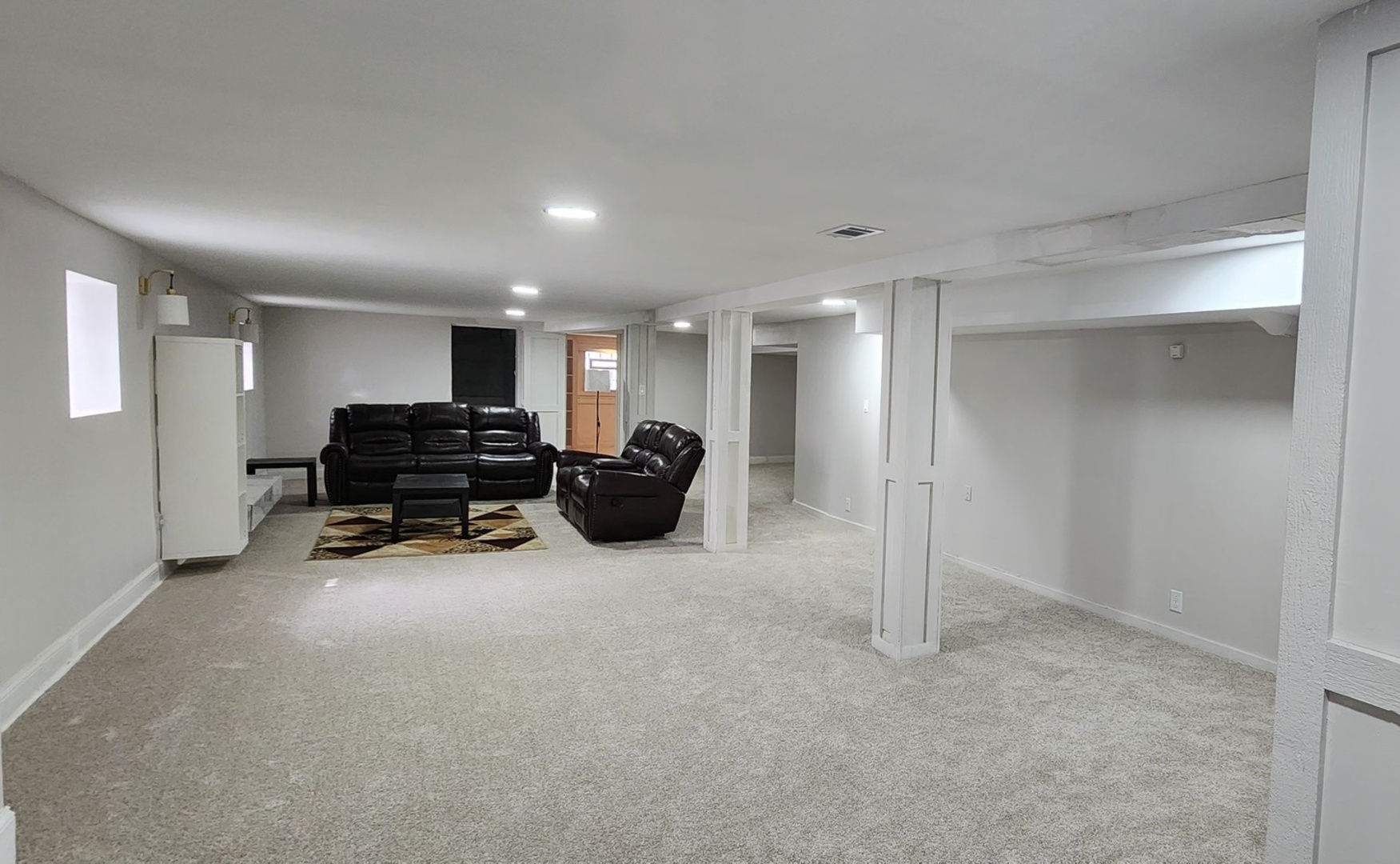 Furnished basement living area with dark leather seating, light carpet, and white support columns.