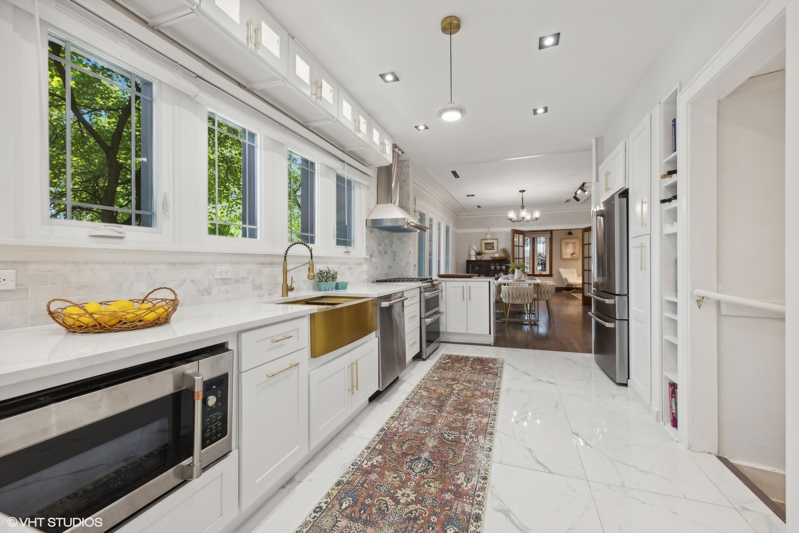 Bright white kitchen with brass farmhouse sink, stainless steel appliances, and large windows overlooking leafy trees.