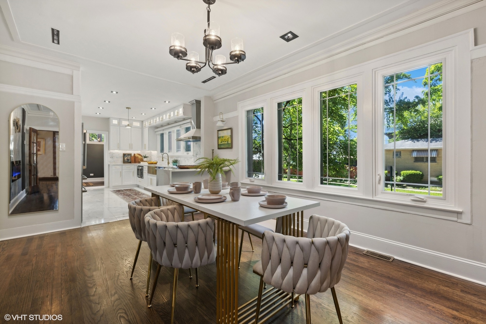 Dining room with a long table, tufted chairs, and large windows beside an open white kitchen.