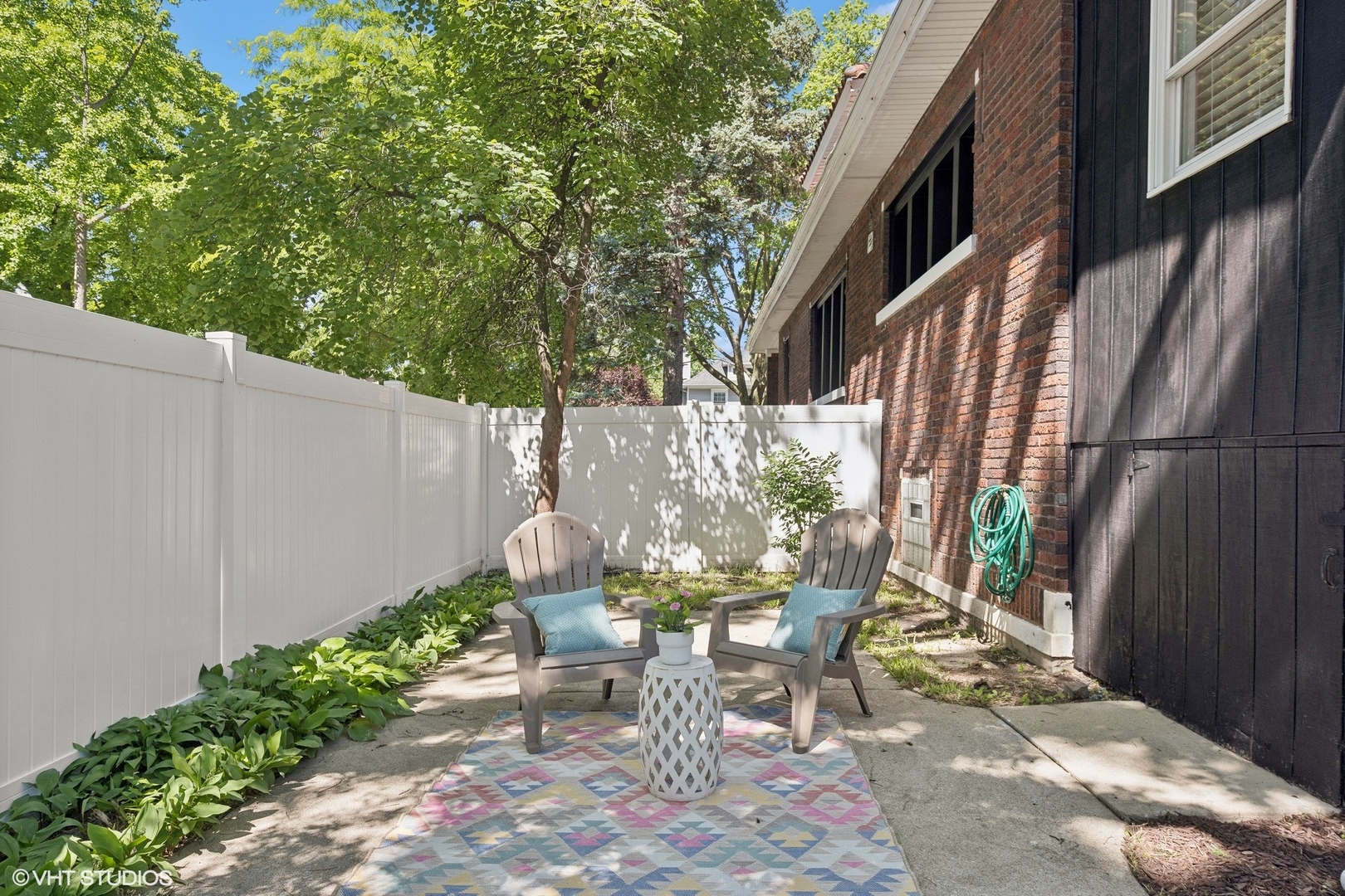 Enclosed patio with two Adirondack chairs, a colorful rug, and a white privacy fence beside a brick house.