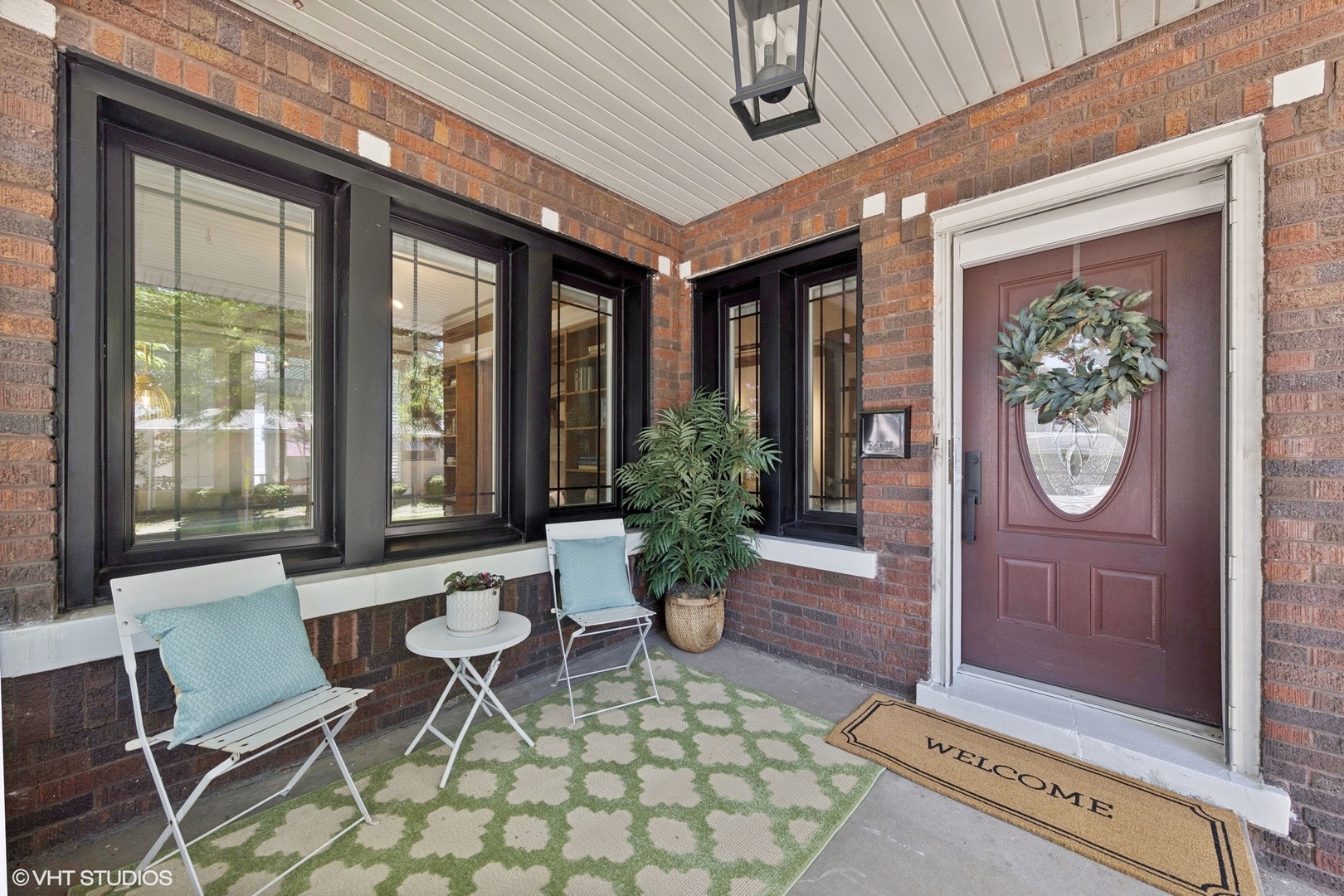 Covered brick front porch with black-framed windows, white chairs, potted plants, and a burgundy front door.