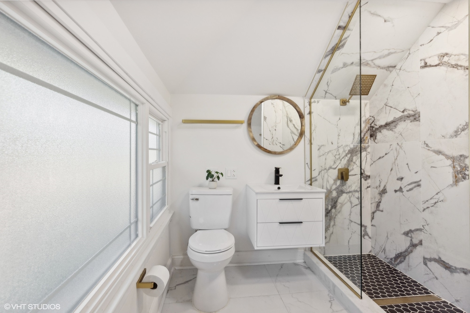 Bathroom with a floating vanity, round mirror, and glass shower with marble-look walls and brass fixtures.