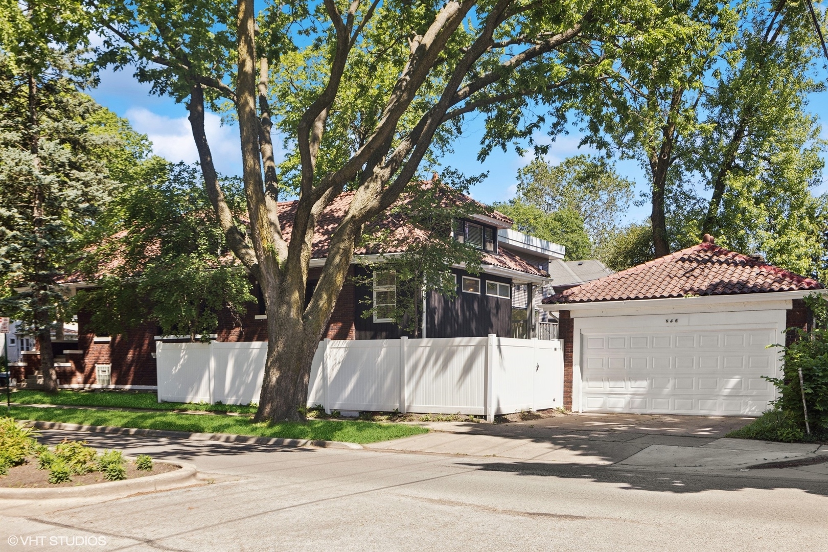 Brick home with a red tile roof, white fence, and attached garage beneath mature trees.