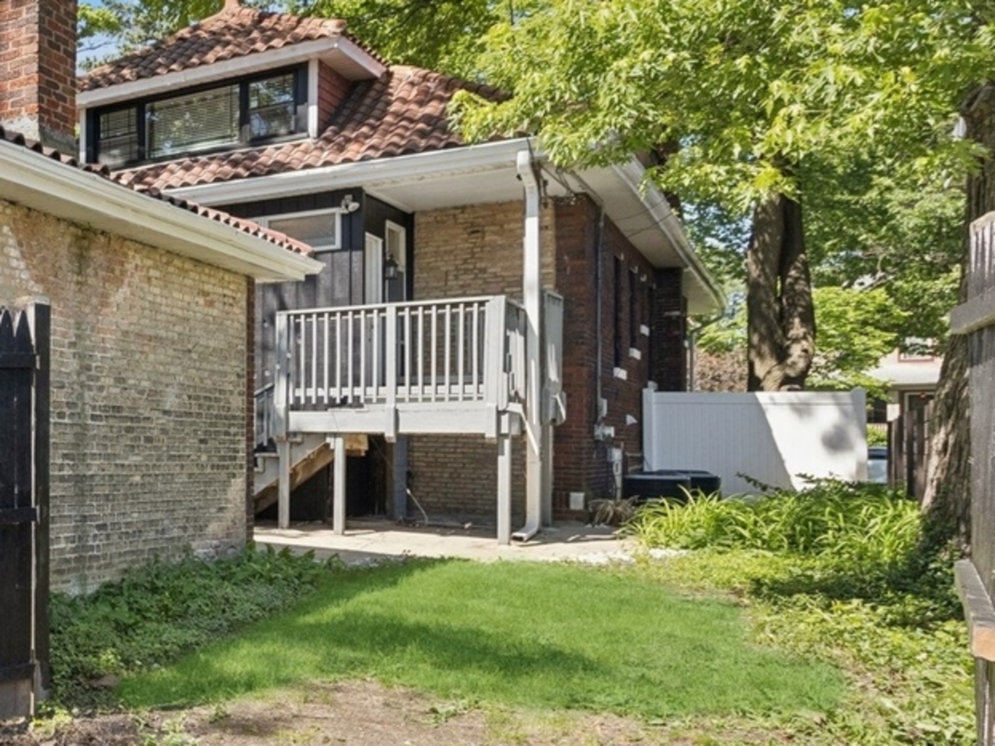 Brick home’s rear exterior with a raised deck and small grassy yard beneath mature trees.