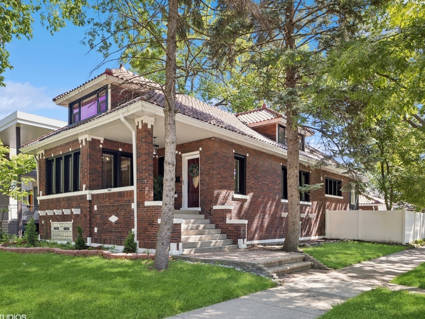 Red brick house with a tiled roof, broad front steps, and mature trees along the sidewalk.