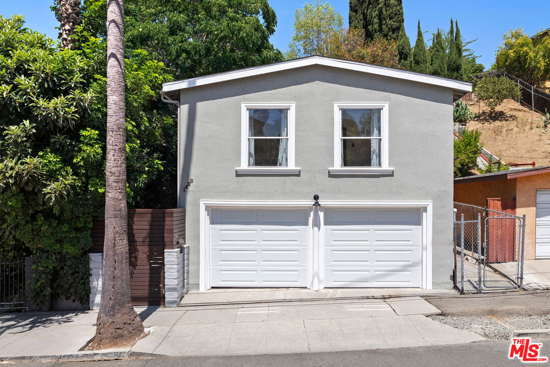 Gray two-story building with twin white garage doors, two upper windows, and a palm tree in front.