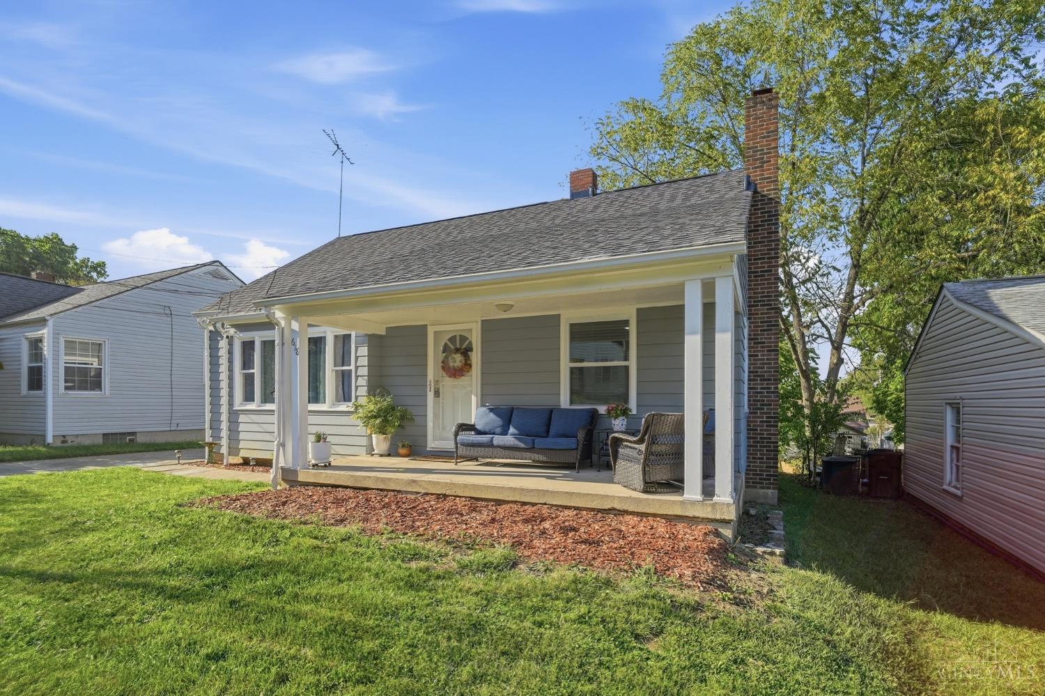 Gray-sided house with a covered porch, blue sofa, brick chimney, and sunny front lawn.