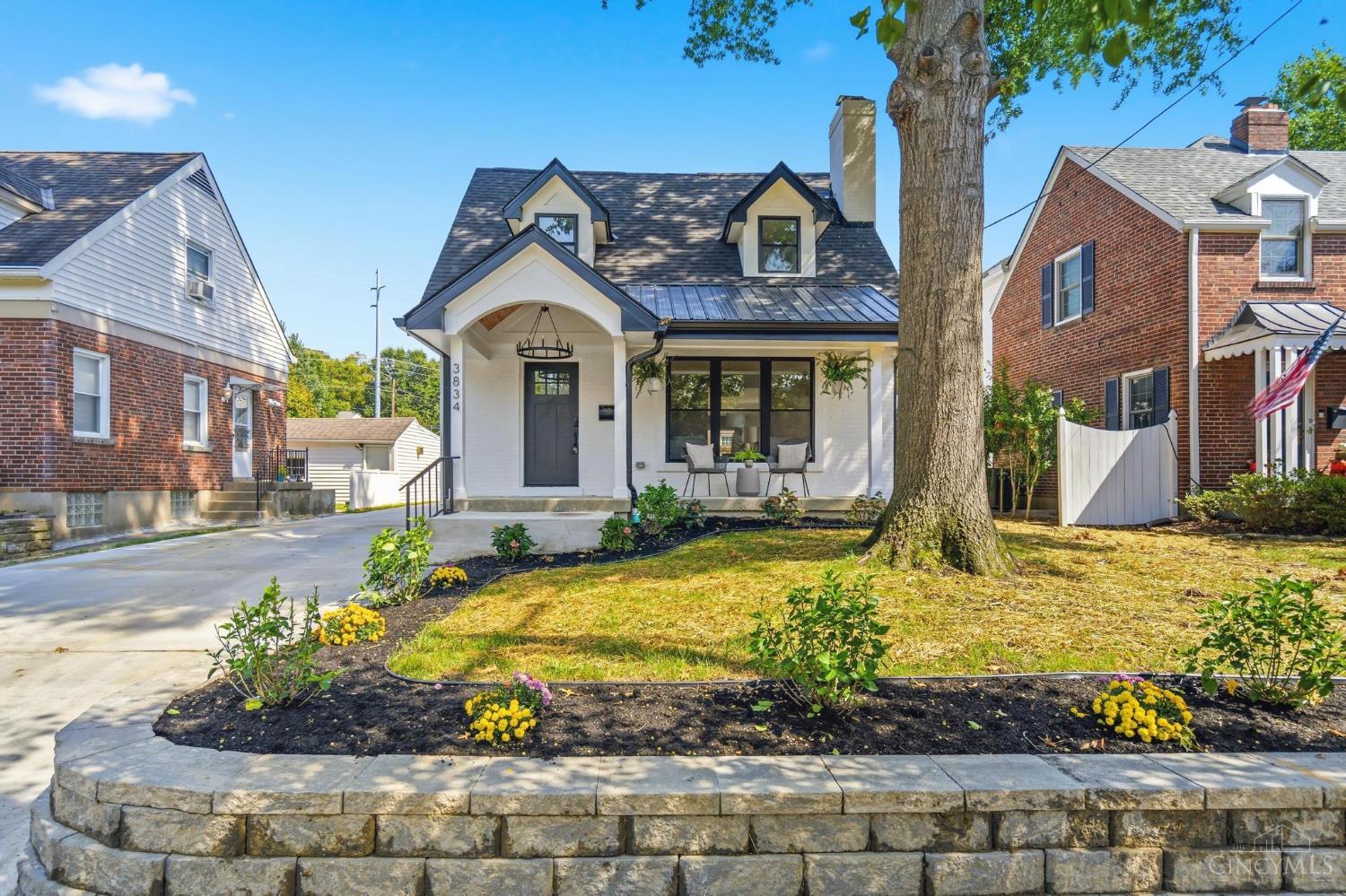 White house with dark trim, two dormers, a covered porch, and a large tree in the front yard.