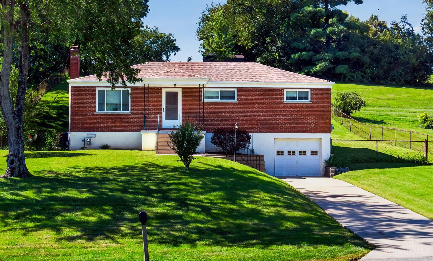 Red brick ranch house with a white garage, curved driveway, and tree-shaded sloping lawn.