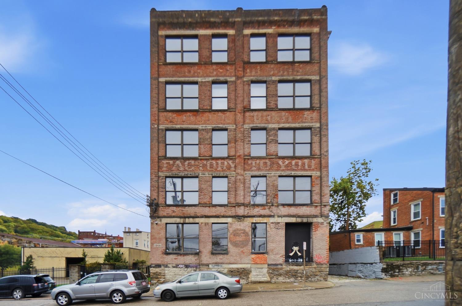 Tall historic brick building with large industrial windows and cars parked along the street.