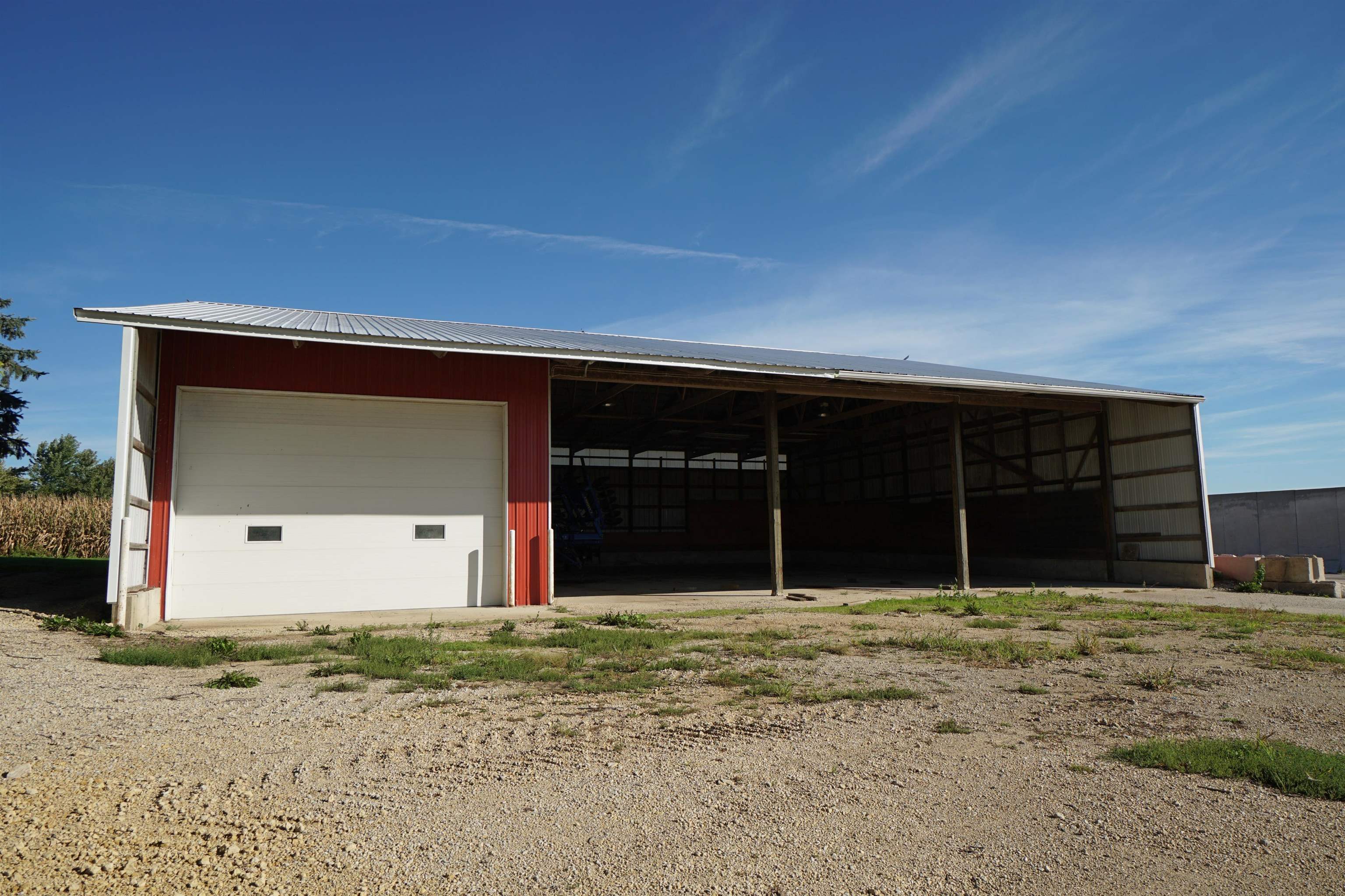 Red agricultural shed with a white garage door and a wide open storage bay beside a gravel lot.