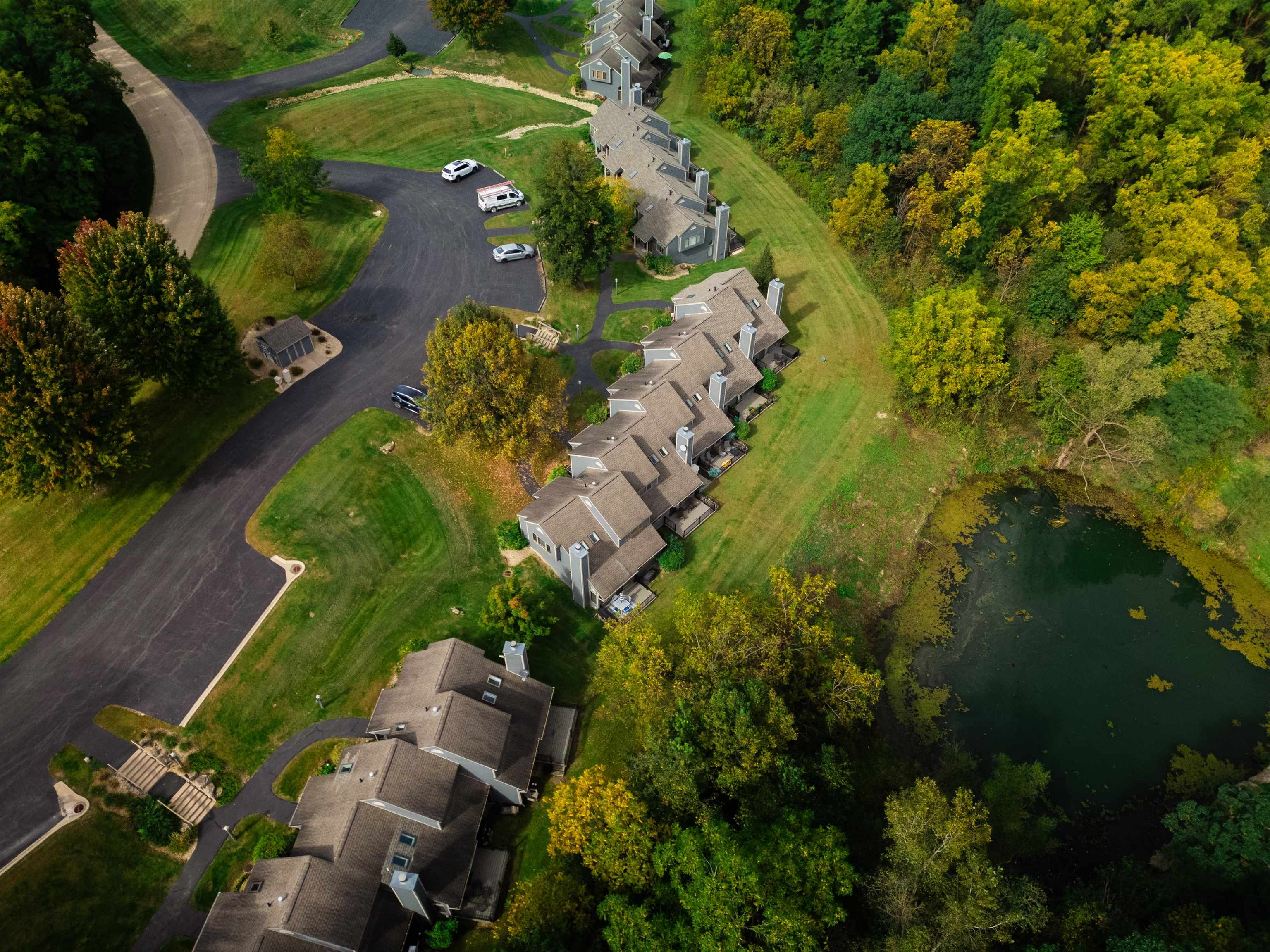Aerial view of attached homes beside a pond, wooded grounds, and a curved drive.