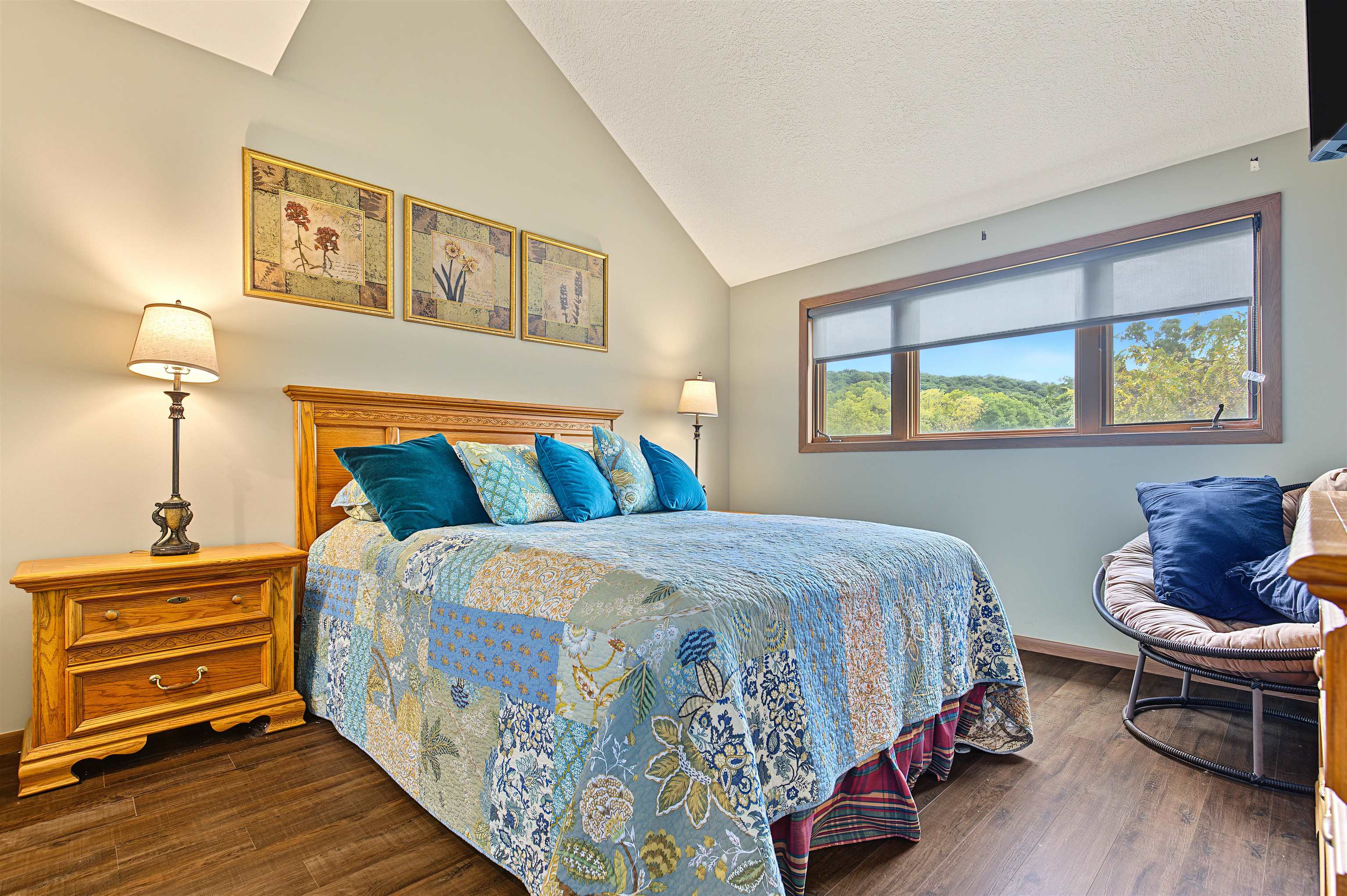 Furnished bedroom with a blue patchwork quilt and a wide window overlooking wooded hills.