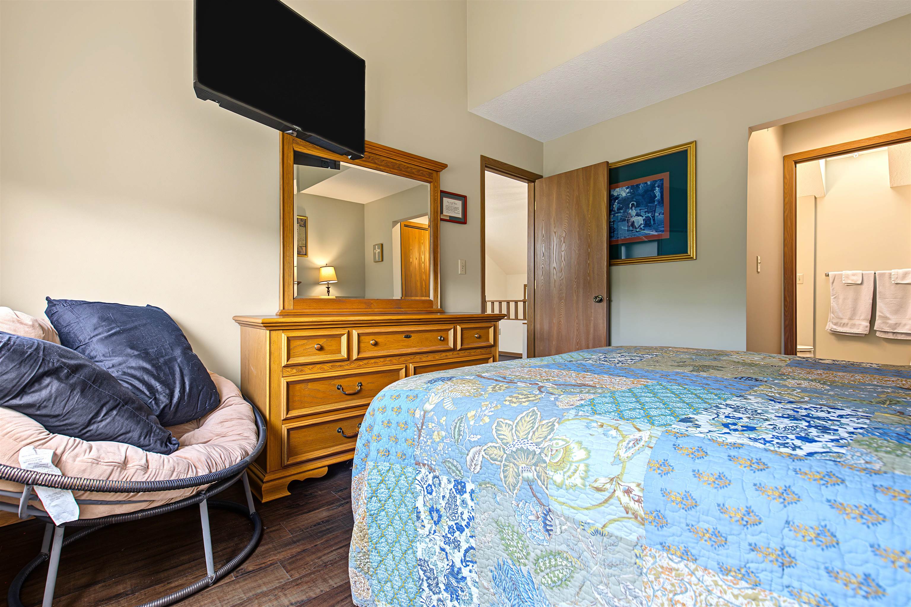Bedroom with a blue floral quilt, wooden dresser and mirror, and cushioned chair.