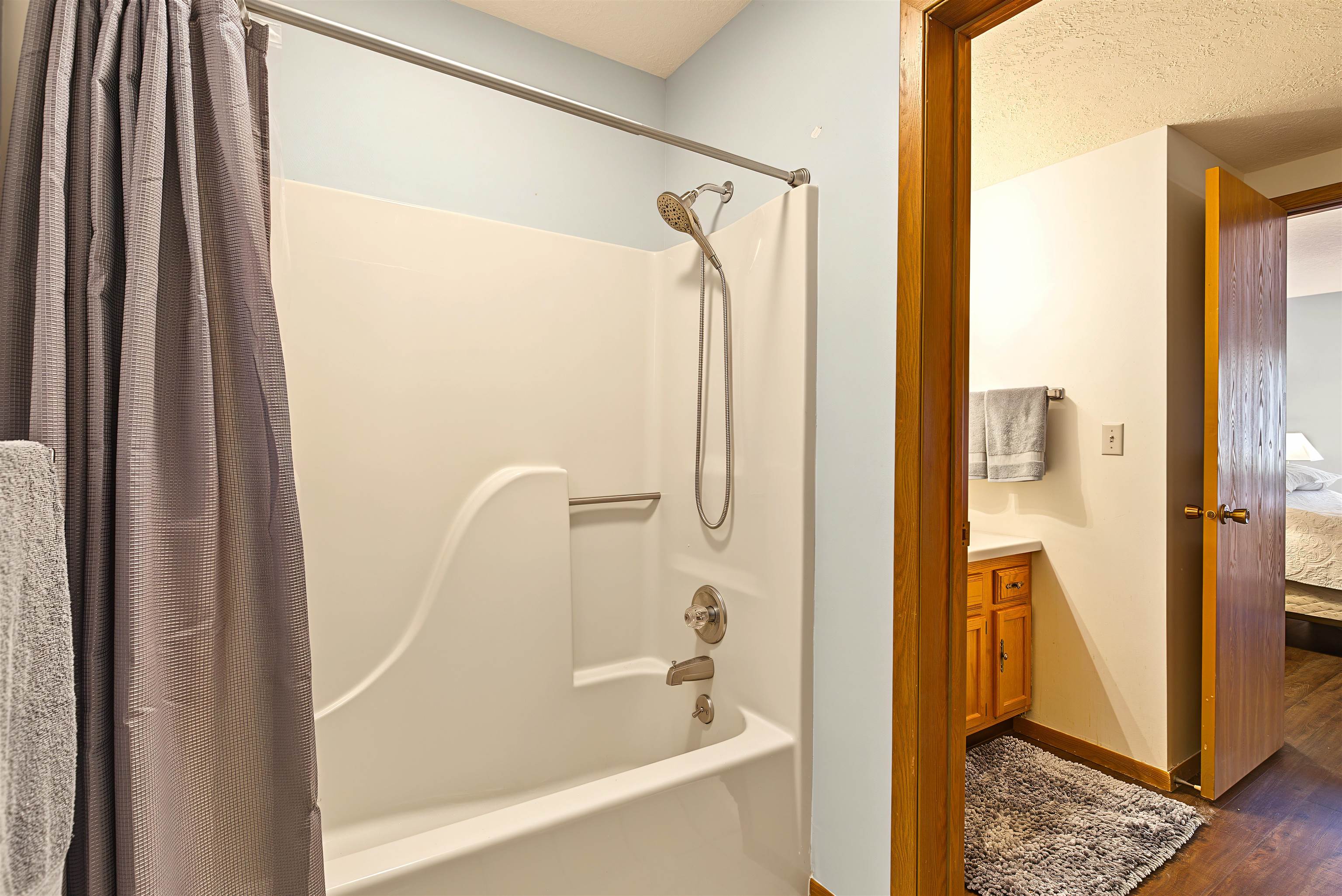 Bathroom with a white tub and shower, gray curtain, and doorway to a wood-cabinet vanity.