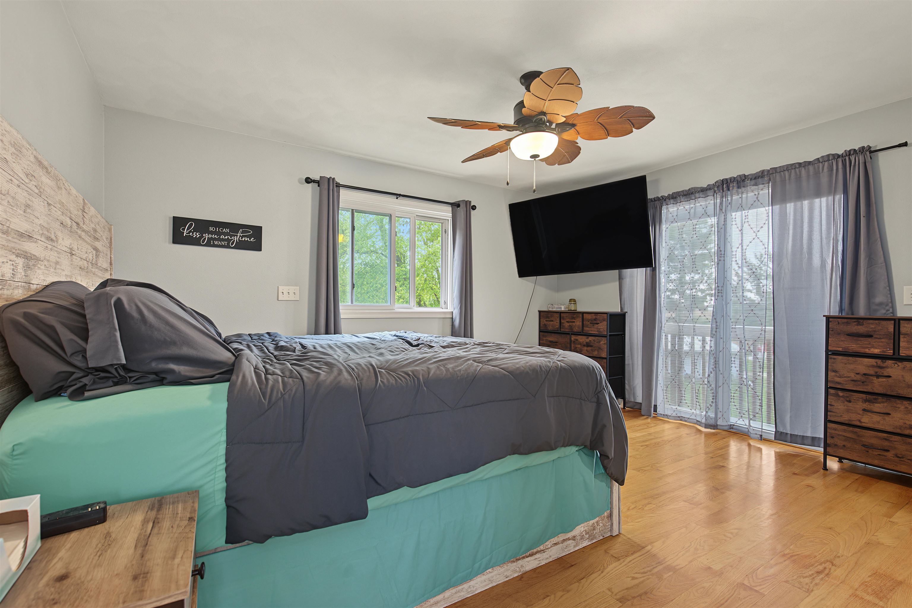 Furnished bedroom with teal and charcoal bedding, hardwood floors, and a wall-mounted television.