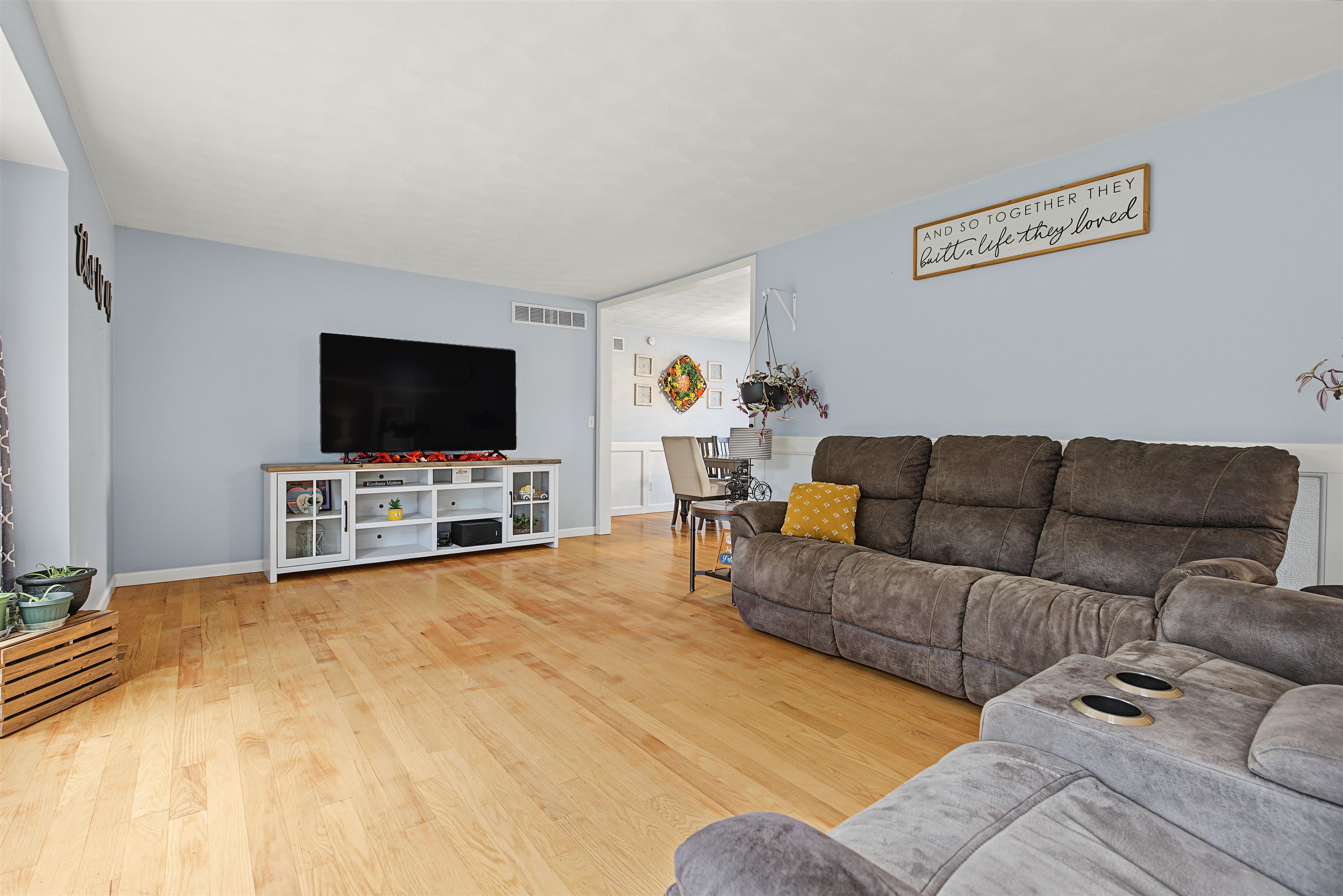 Living room with a gray sectional, television, pale blue walls, and light wood flooring; dining area beyond.