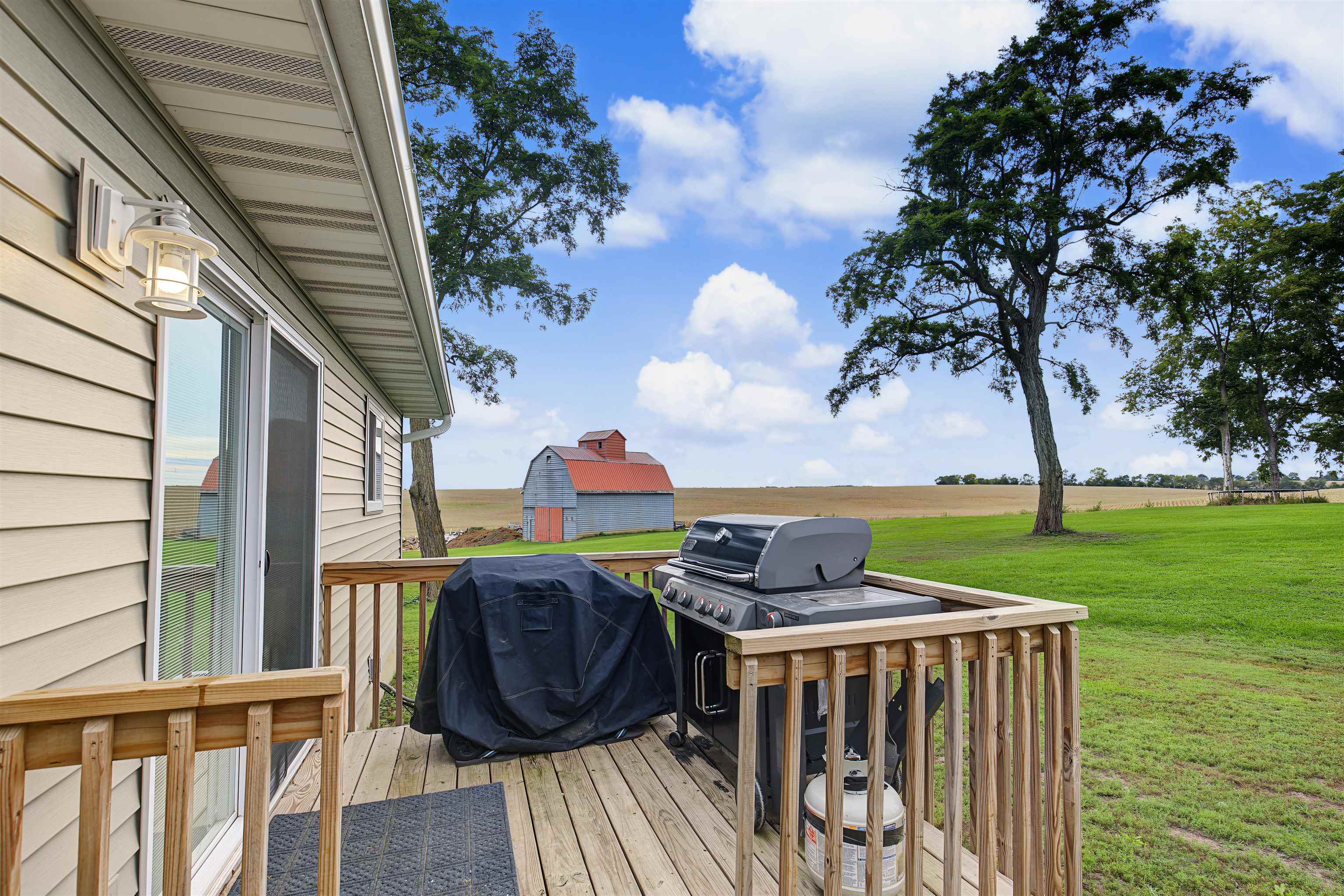 Wooden deck with a gas grill overlooks a green lawn, mature trees, farmland, and a red-roofed barn.
