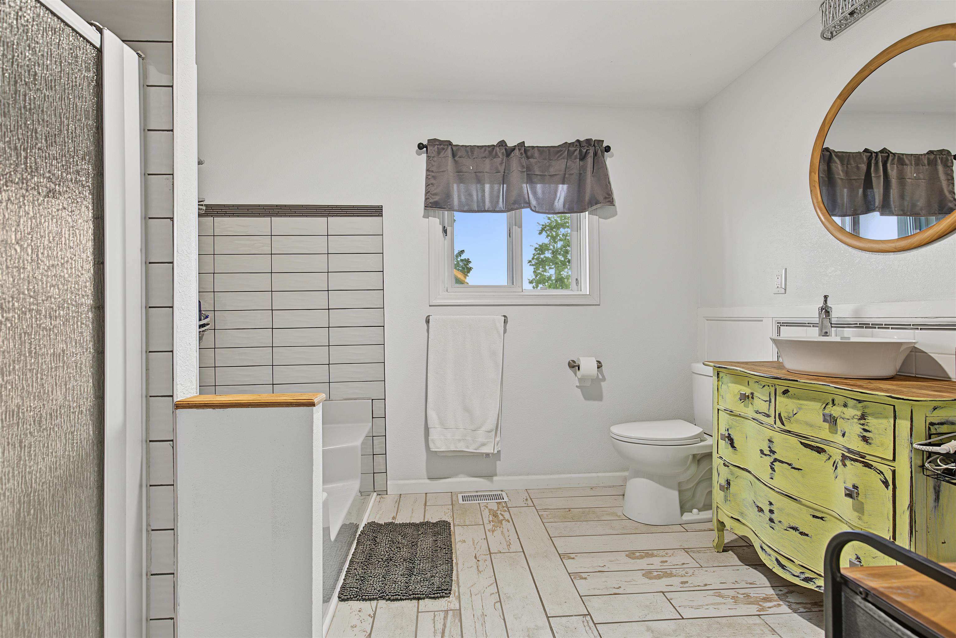 Bathroom with a tiled shower and a distressed yellow-green vanity beneath a round wood-framed mirror.