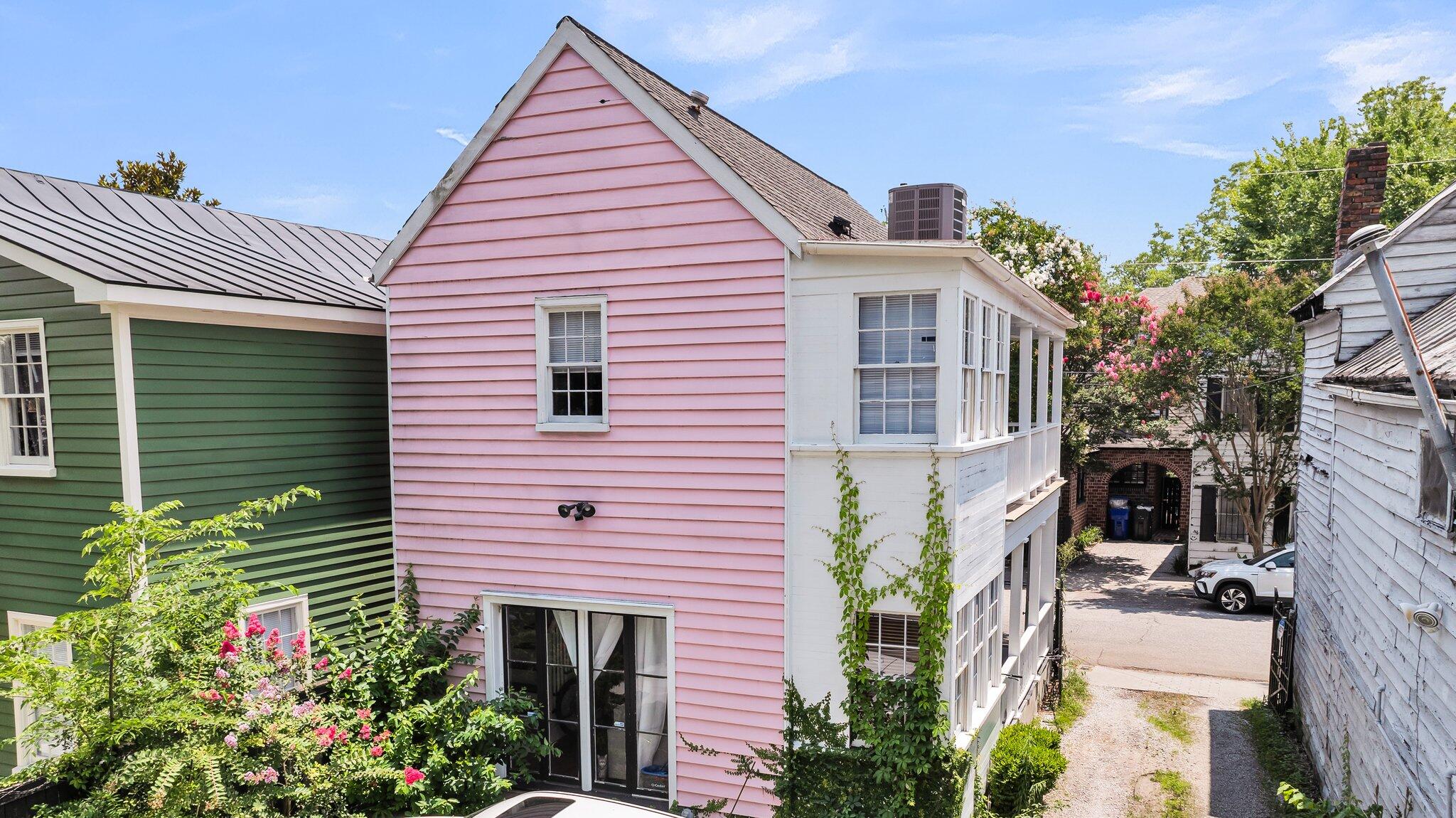 Pink clapboard house with a steep gable, white enclosed porch, and flowering trees beside a narrow driveway.