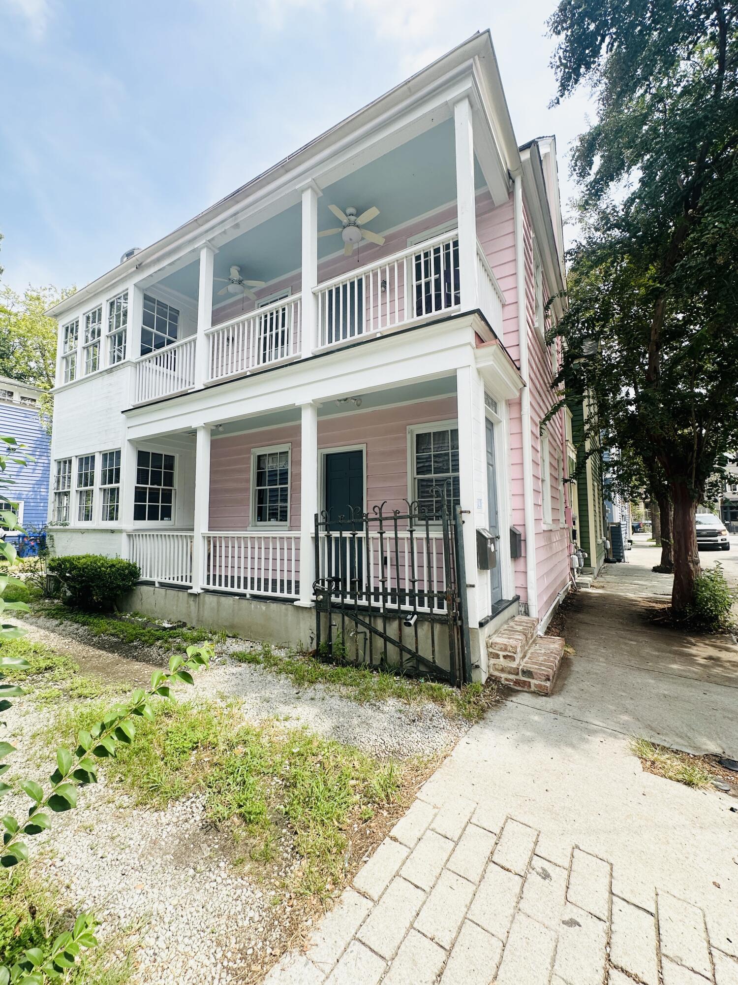 Pink two-story house with white-trimmed porches and an upper balcony beside a tree-lined sidewalk.