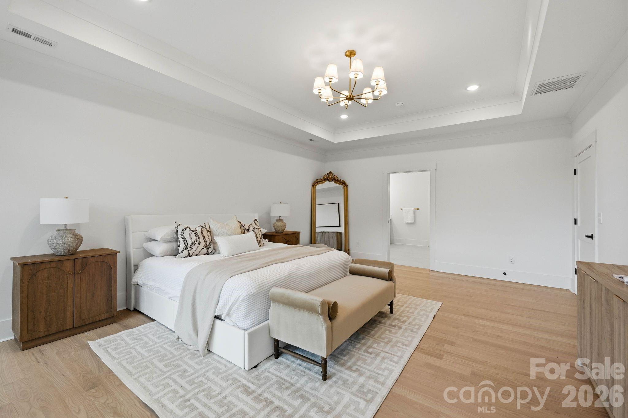 Staged bedroom with a white bed, patterned rug, wood nightstands, and a brass chandelier.