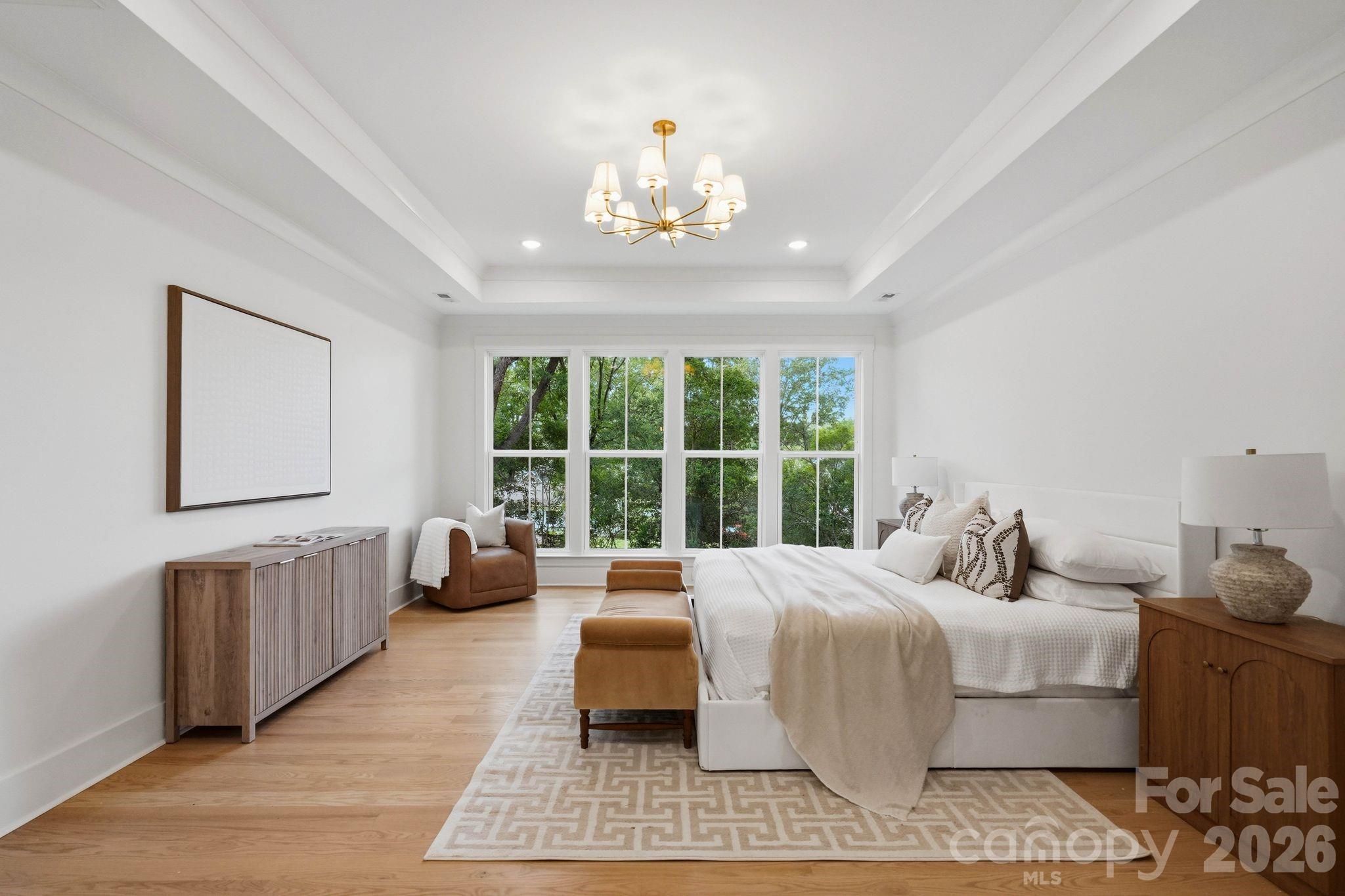Staged bedroom with a white bed, brass chandelier, and large windows overlooking leafy trees.