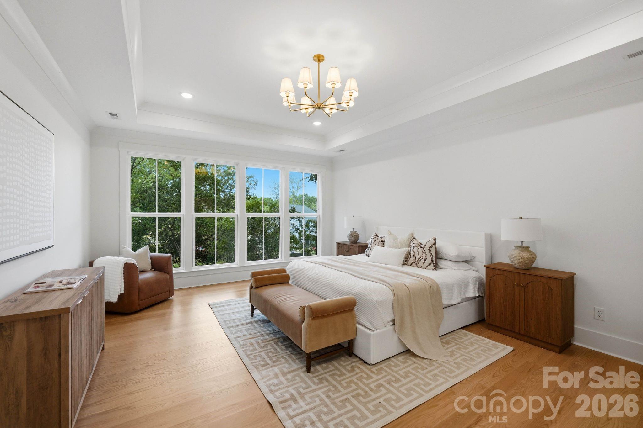 Bright staged bedroom with a white bed, tan bench, and large windows overlooking trees.