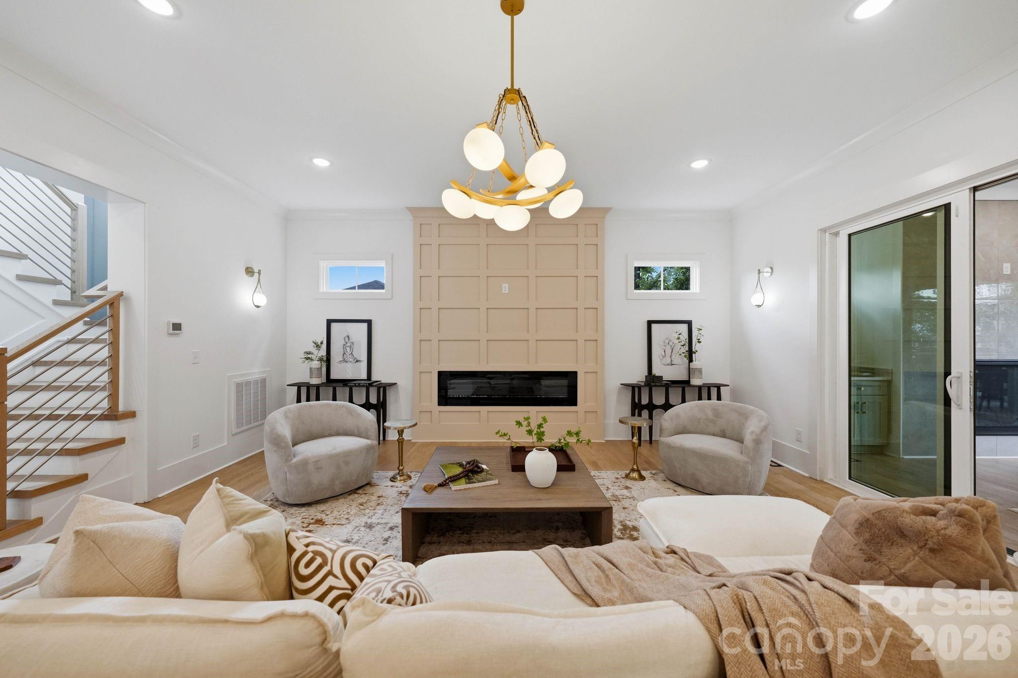 Staged living room with a paneled fireplace, sculptural chandelier, and staircase.