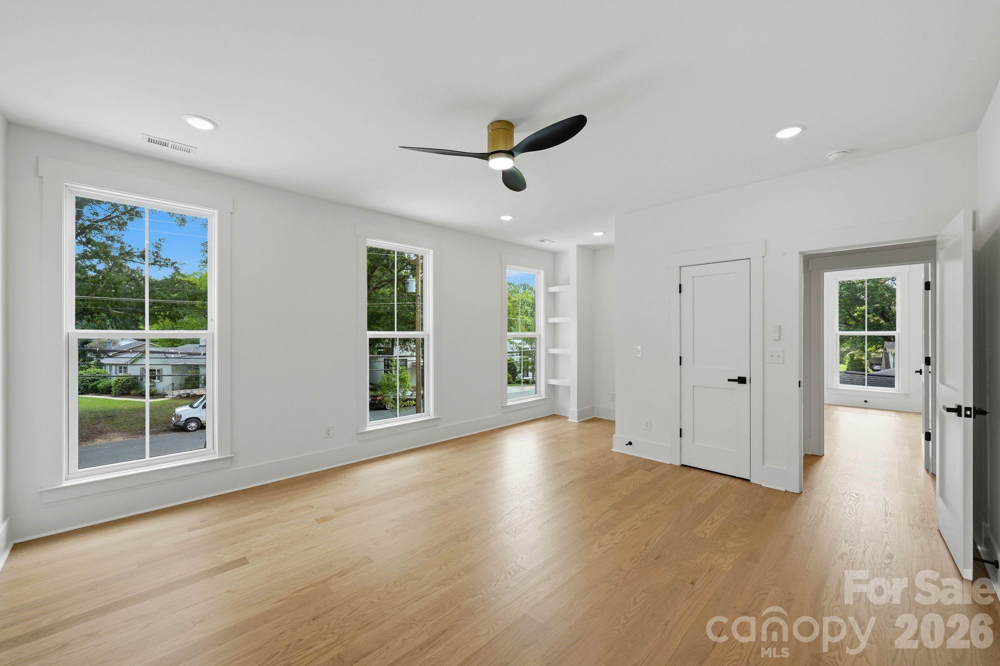 Vacant room with light hardwood floors, a black ceiling fan, and large windows overlooking trees.