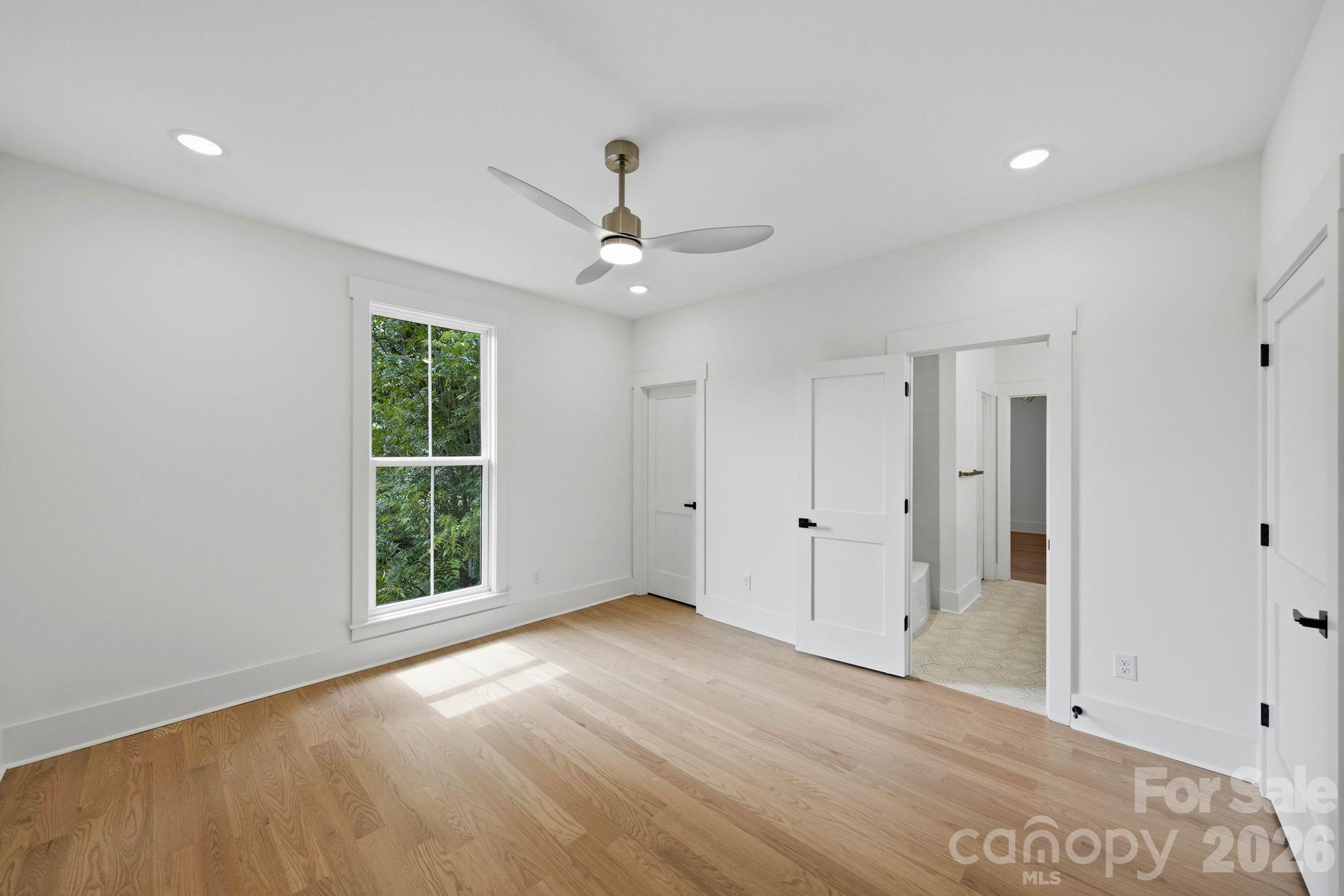 Empty bedroom with light hardwood floors, a ceiling fan, and a large window overlooking greenery.