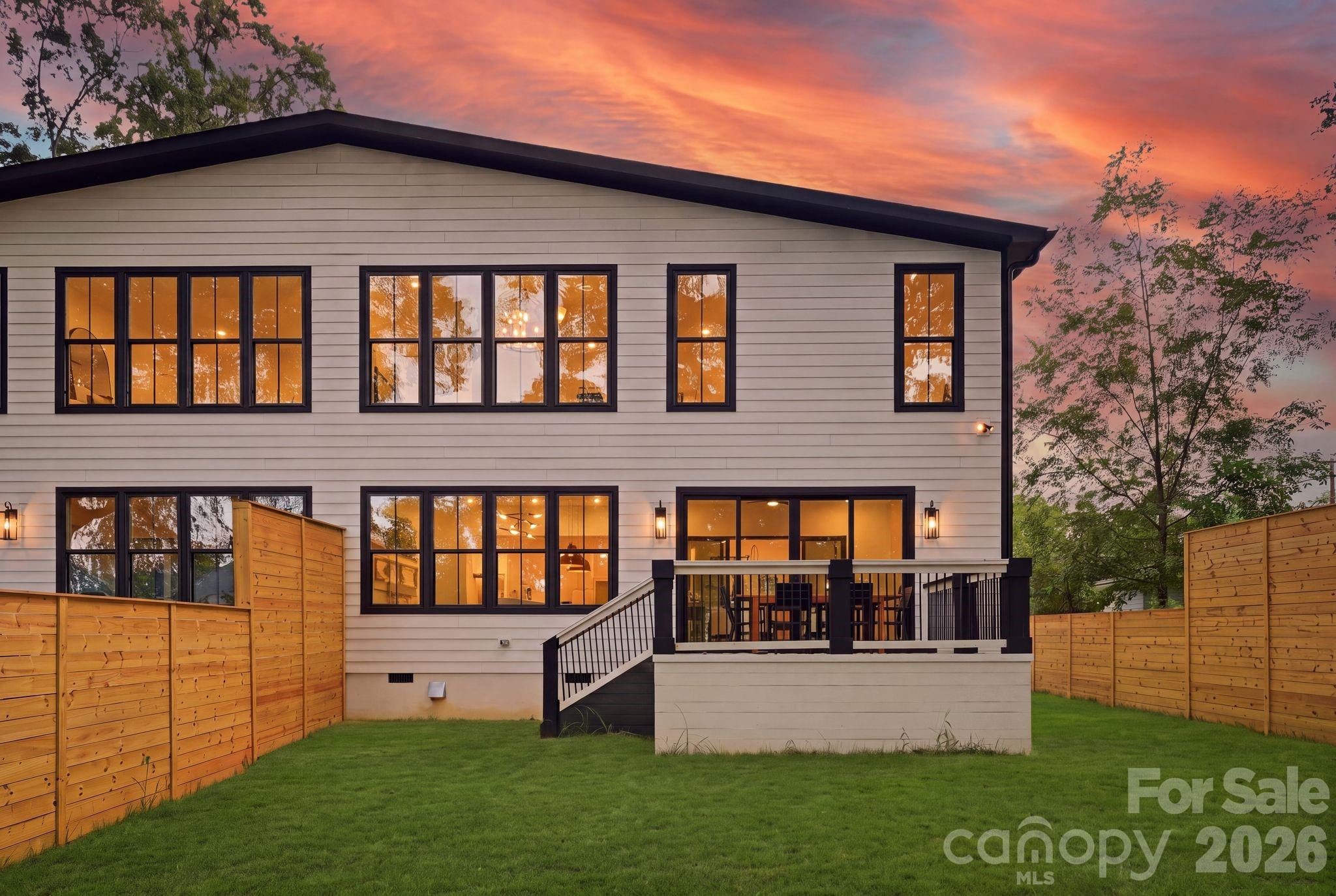 Two-story home’s rear exterior with a raised deck, fenced lawn, and warmly lit windows at dusk.