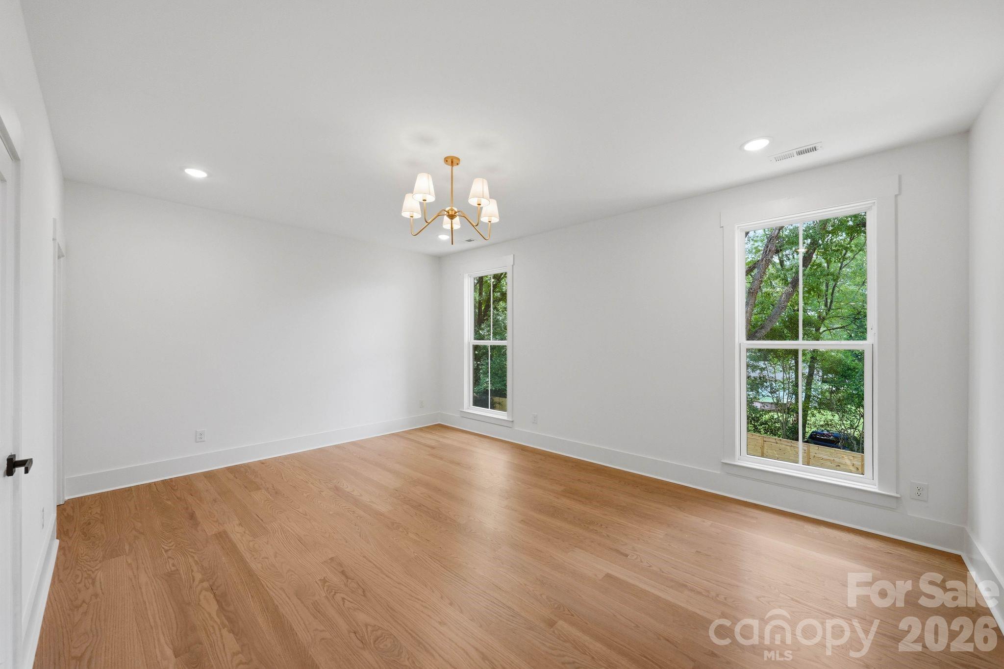 Empty room with hardwood floors, white walls, two windows overlooking trees, and a brass chandelier.