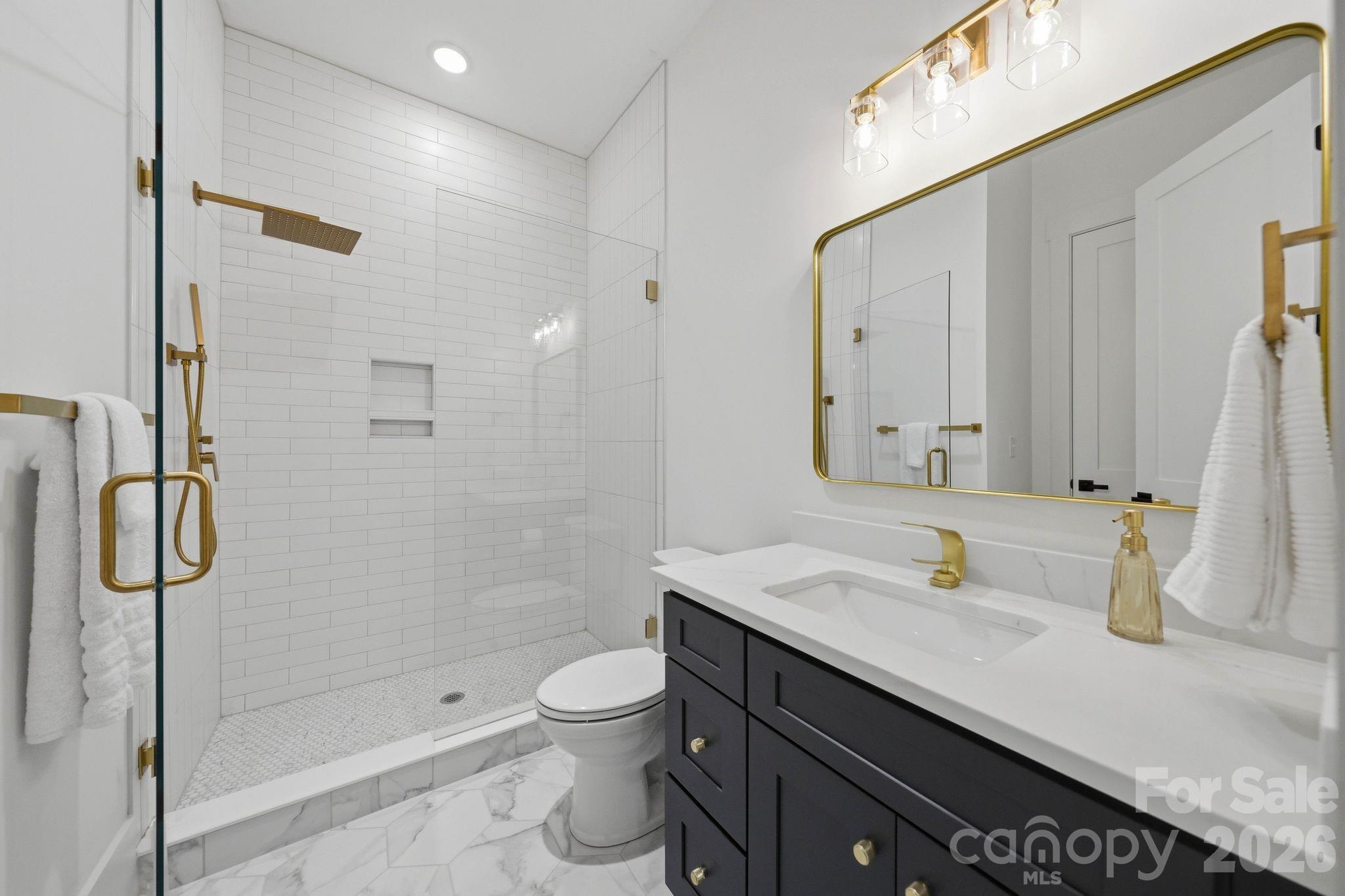 Modern bathroom with a glass walk-in shower, white tile, dark vanity, and brass fixtures.