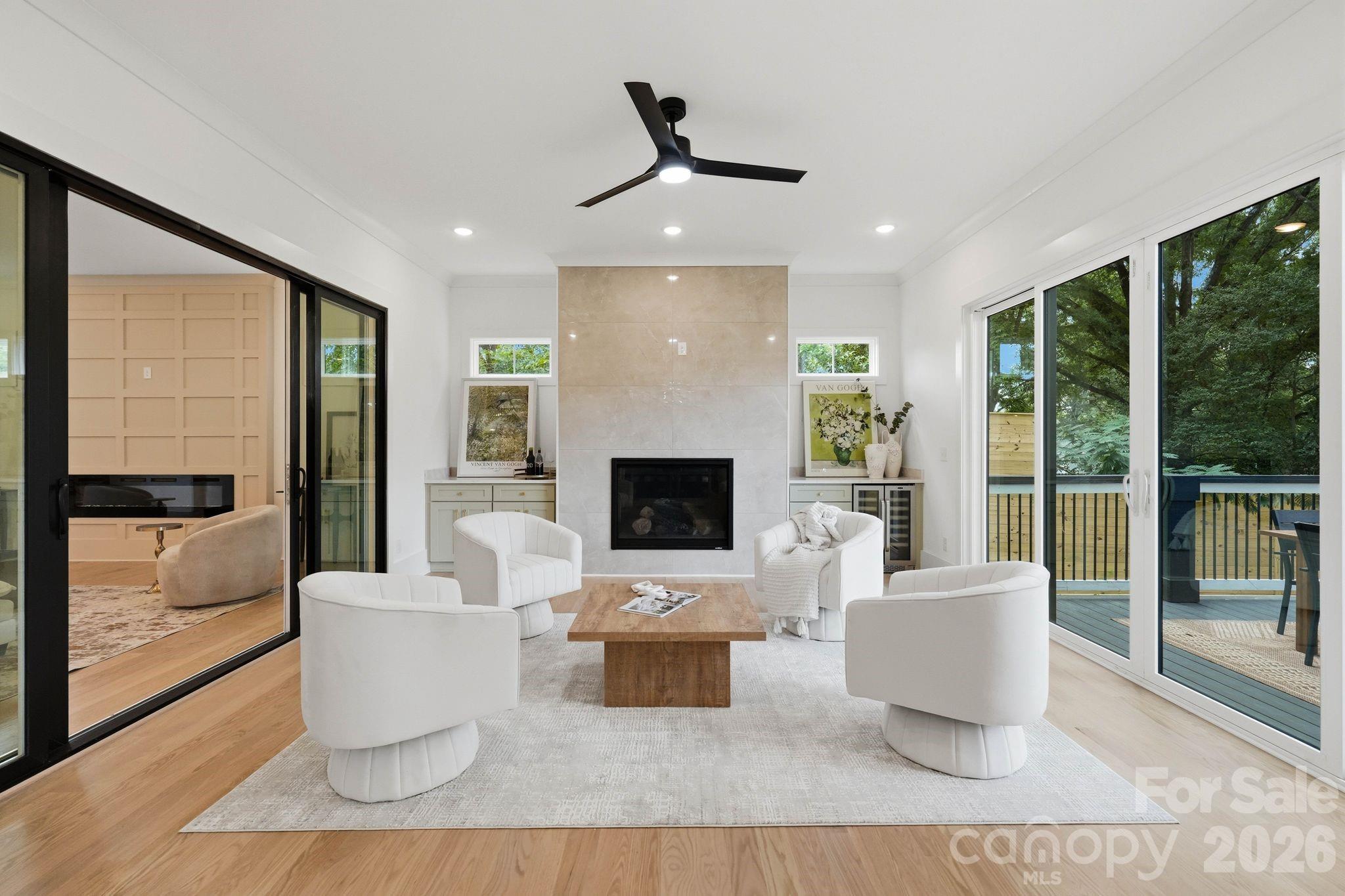 Staged living room with a tiled fireplace, white chairs, and sliding doors overlooking a leafy deck.